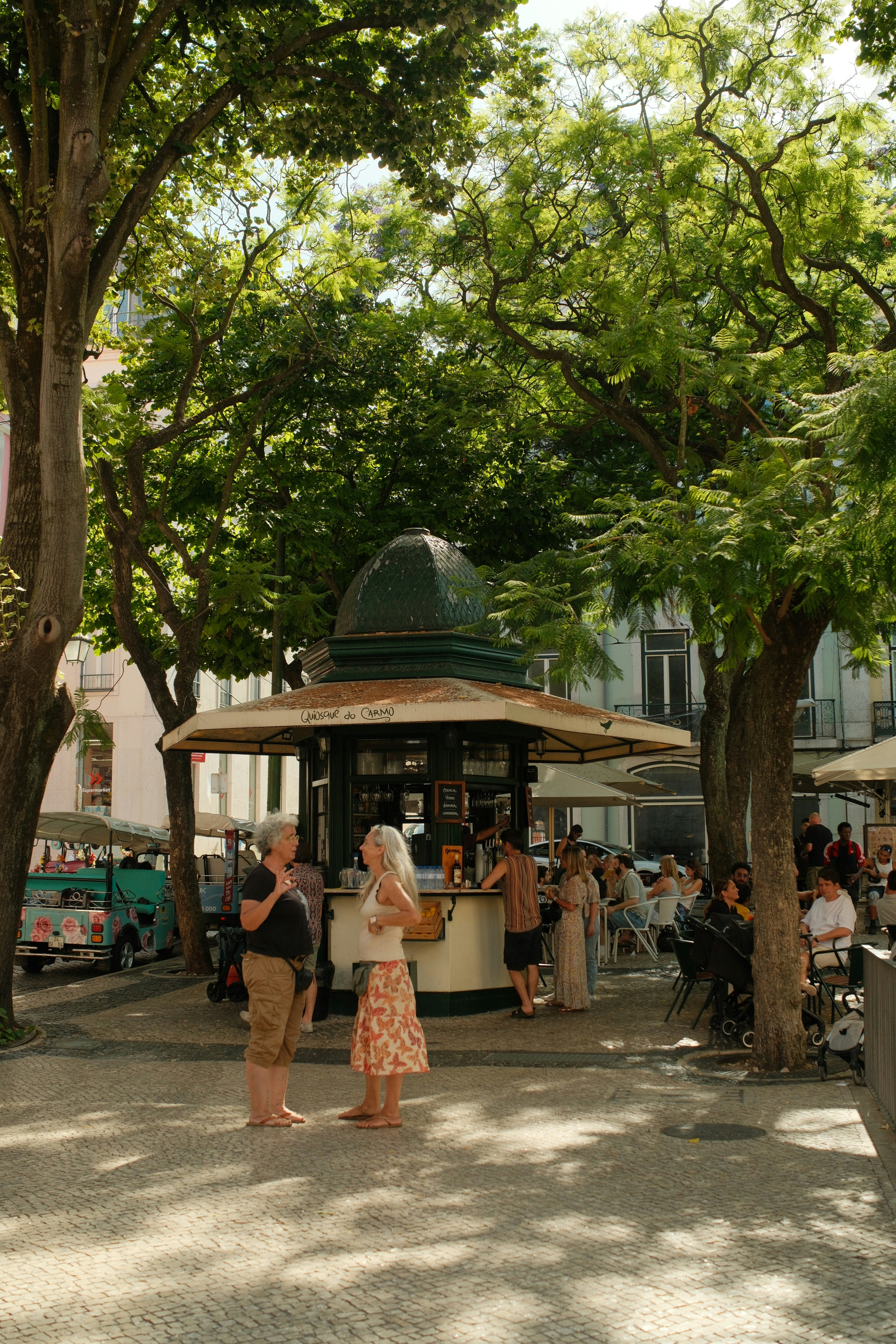 Two women talking in front of a kiosk in a tree-lined Lisbon square