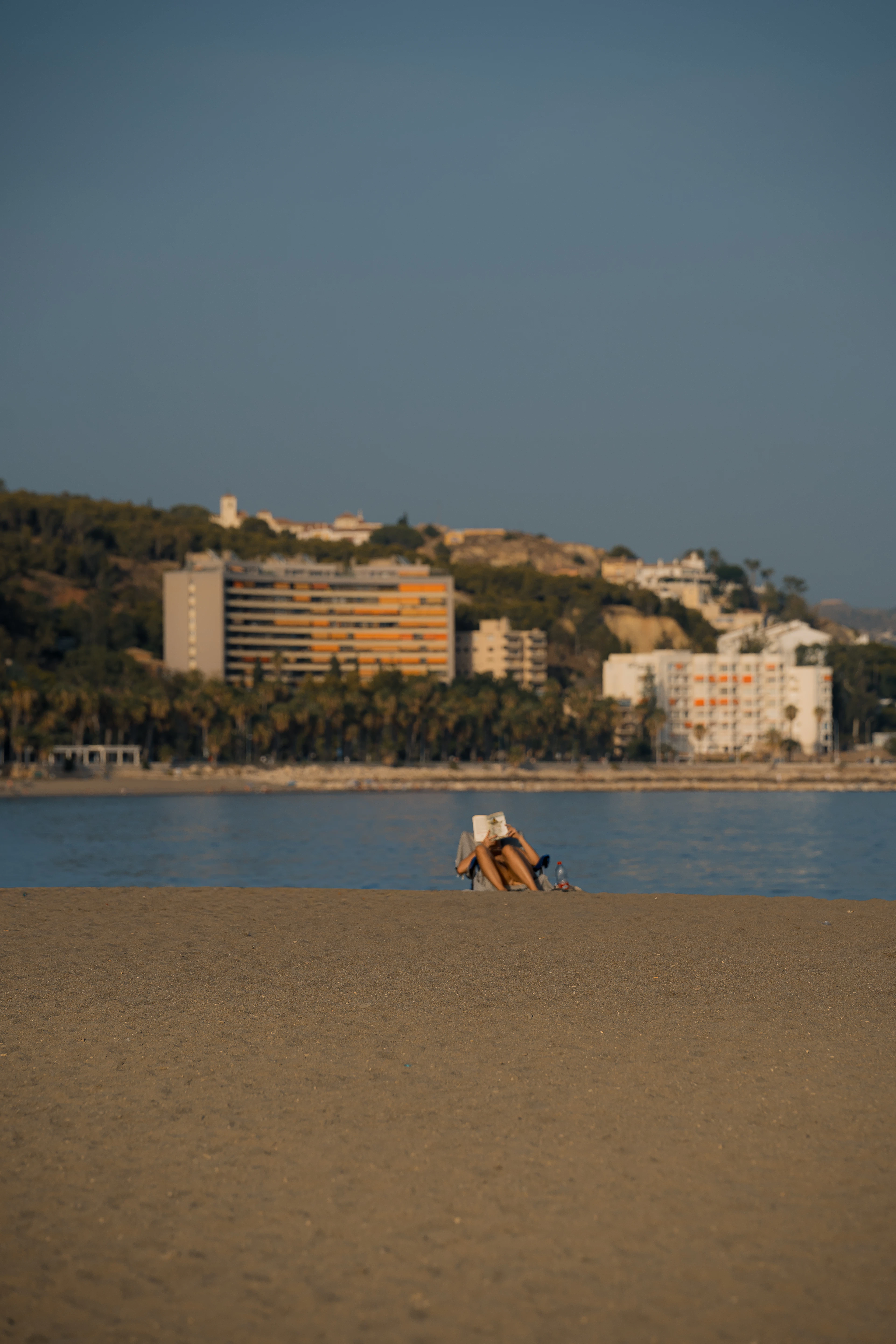 A person reads on a Málaga beach at golden hour with the city's hillside skyline behind.