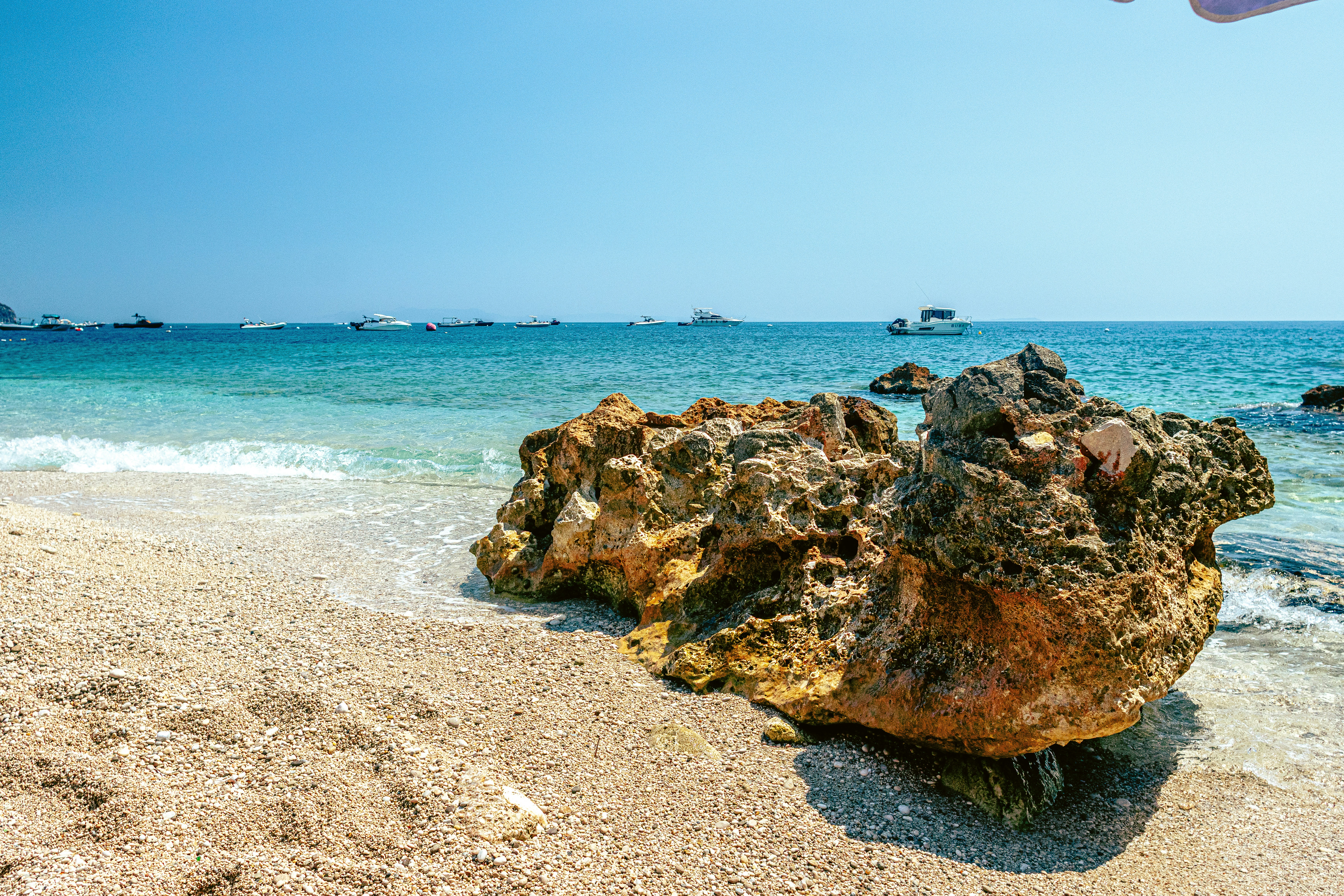 A large porous rock on a pebble beach with boats on the horizon