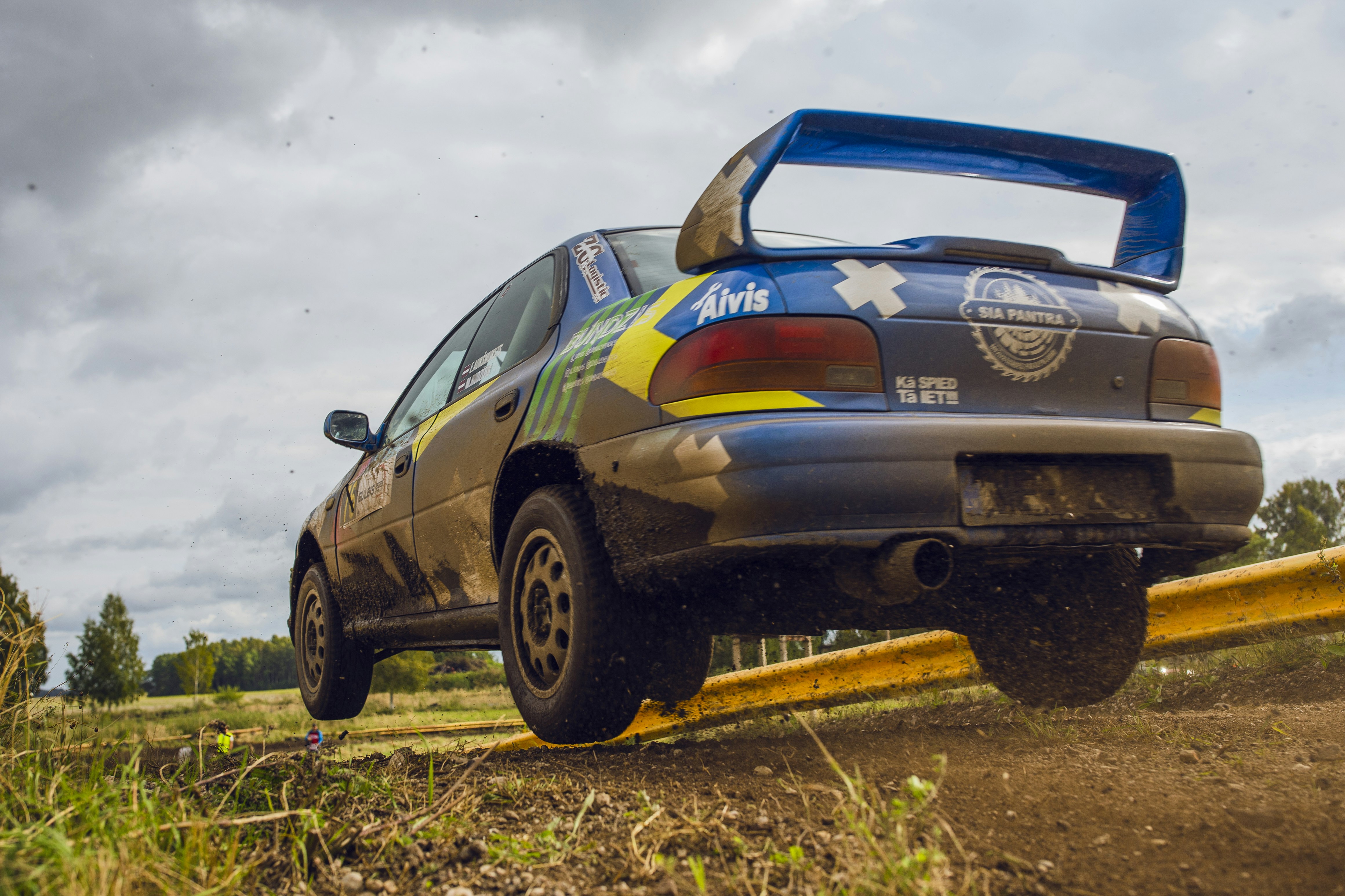 A blue rally car jumping over a yellow barrier on a dirt track