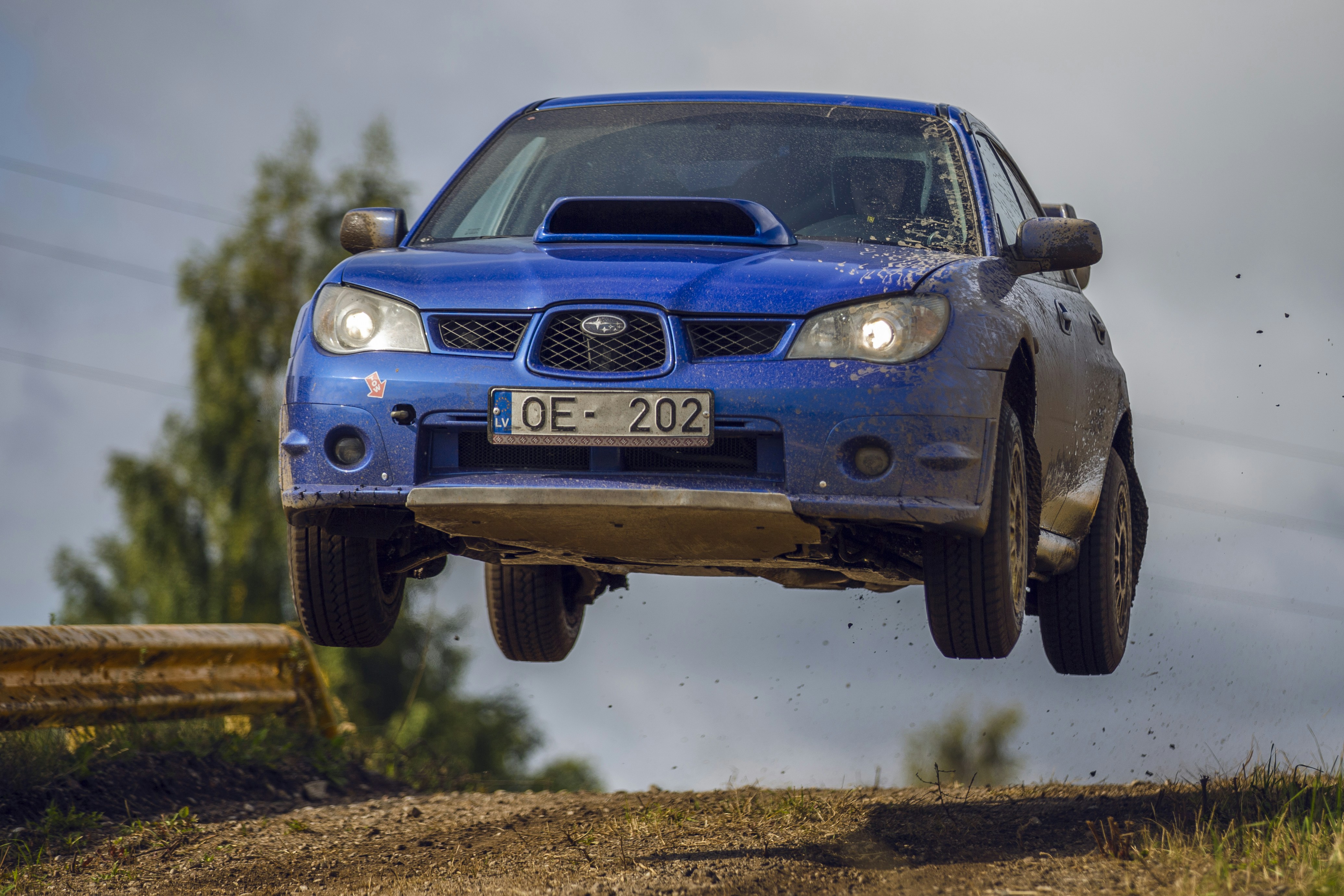 A blue rally car jumping over a dirt track with trees in the background