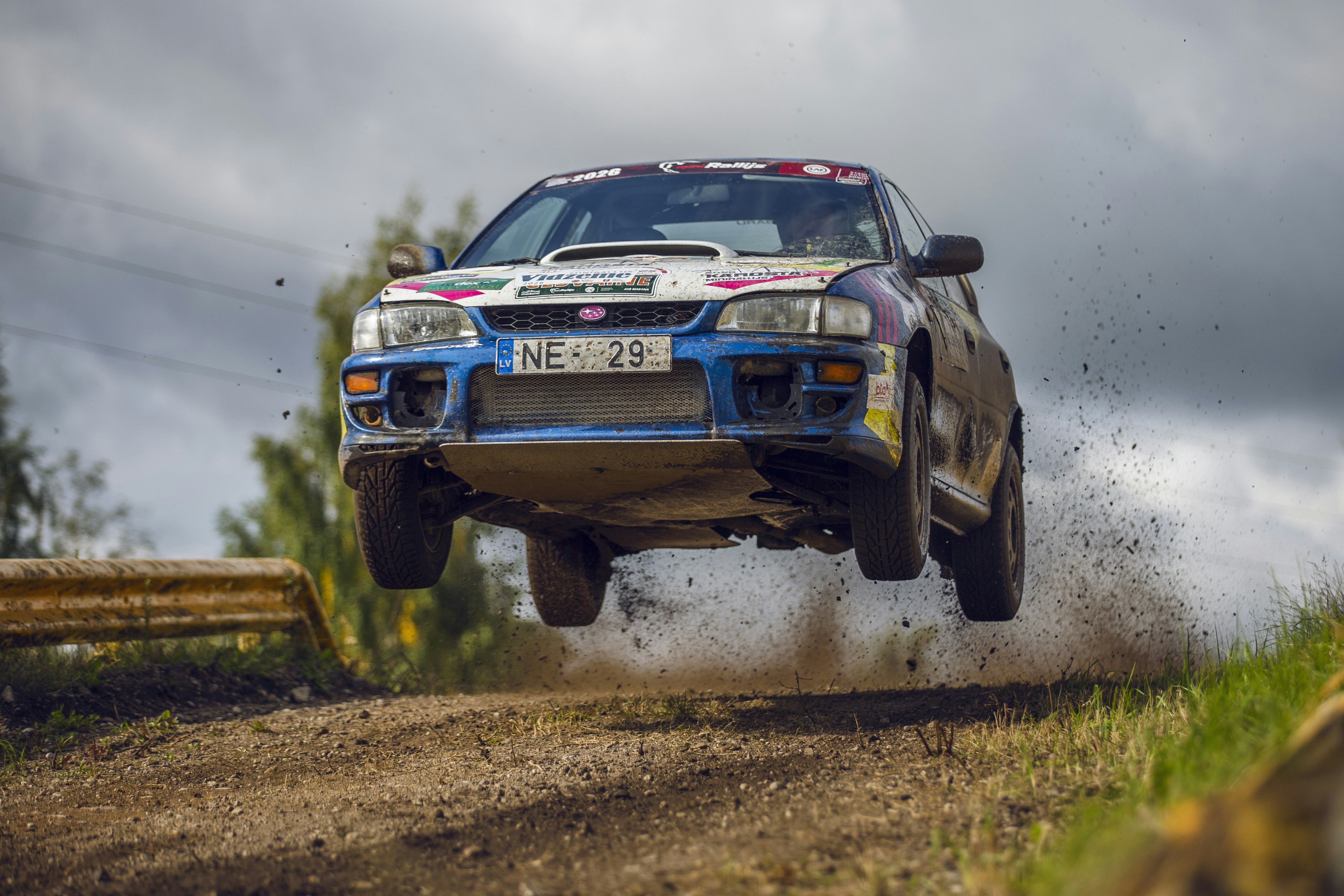 A blue rally car jumping over a dirt track with mud spraying