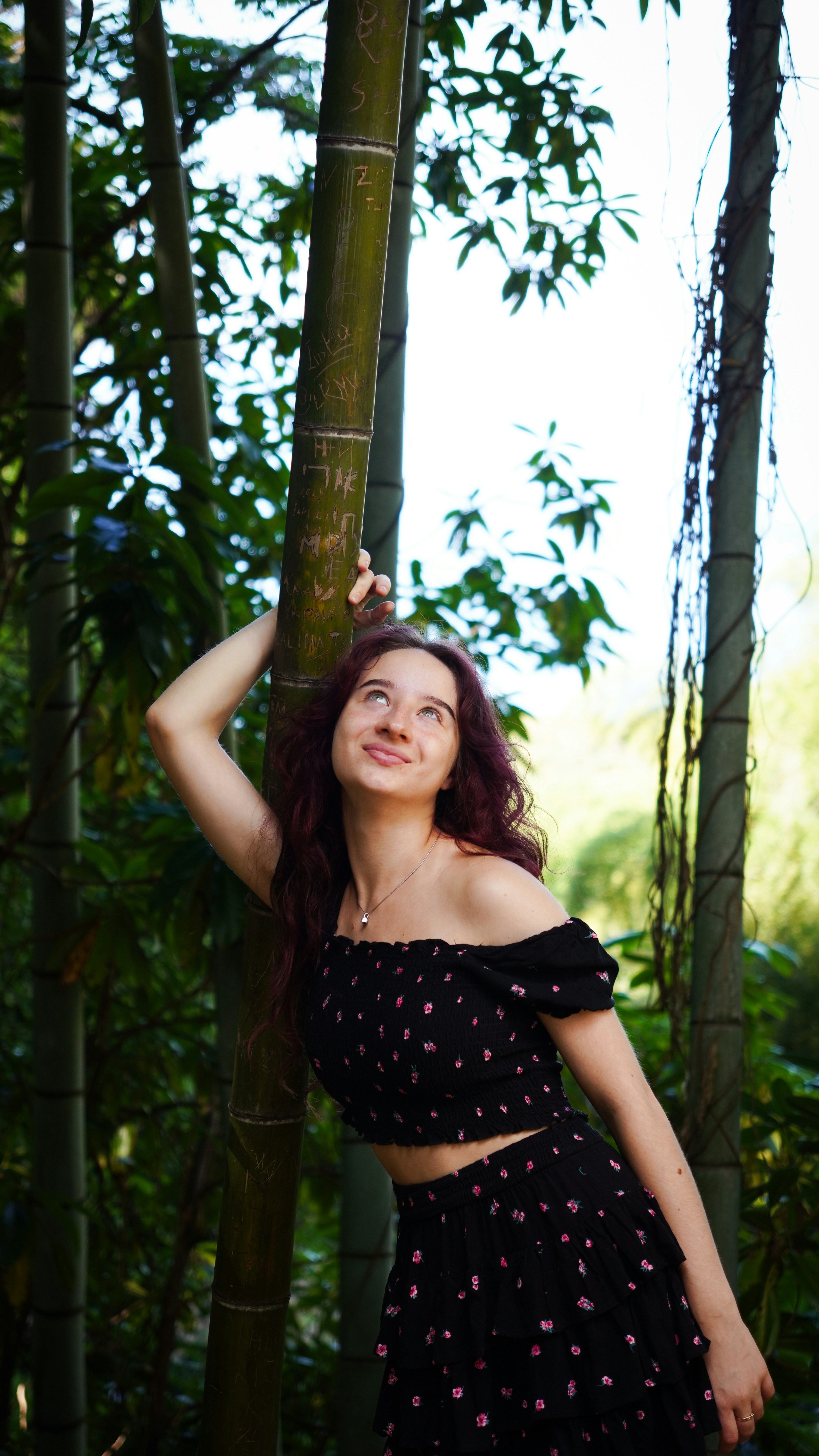 A woman with long dark hair leaning against a bamboo stalk in a forest