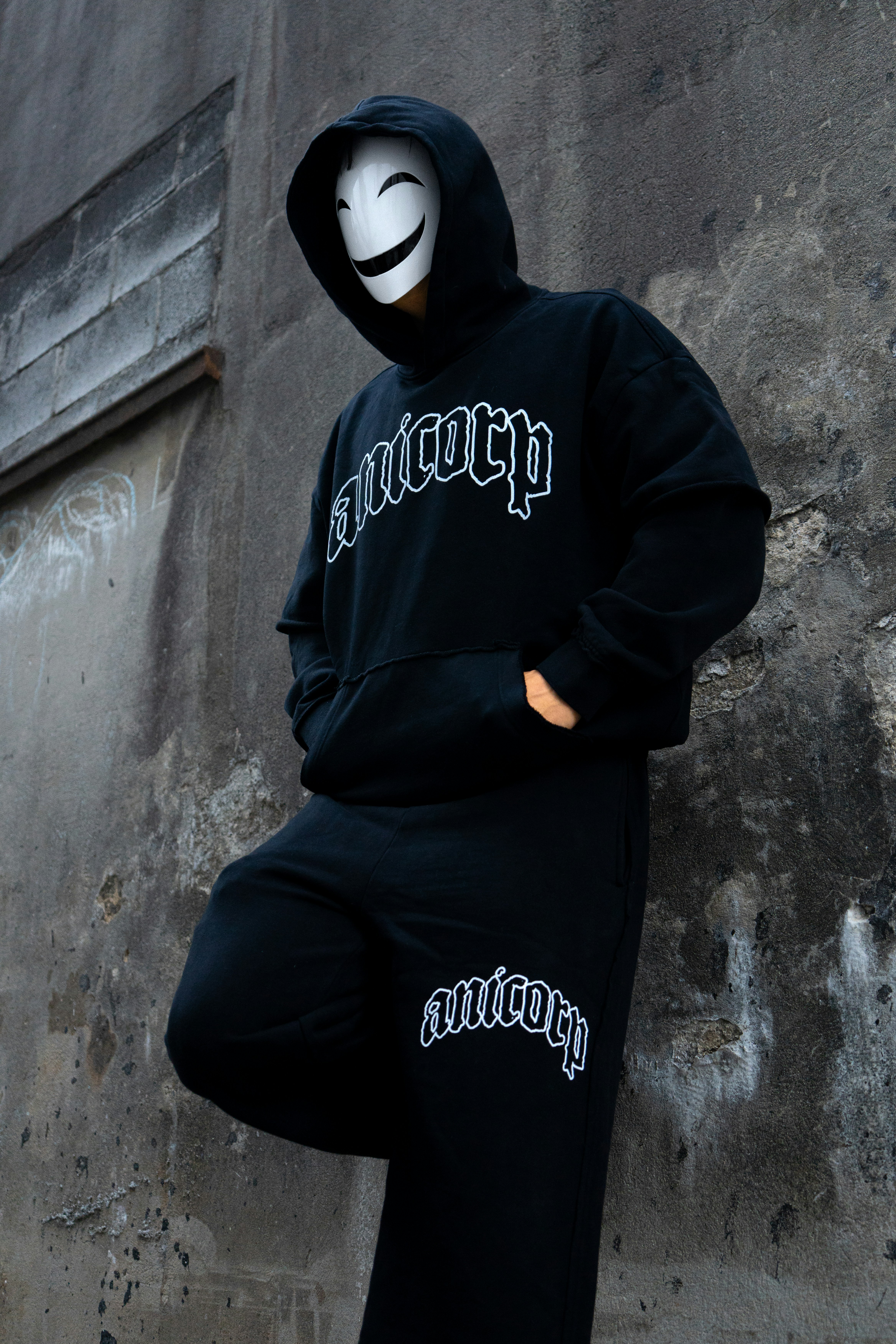 A person in a black hoodie and mask leaning against a concrete wall