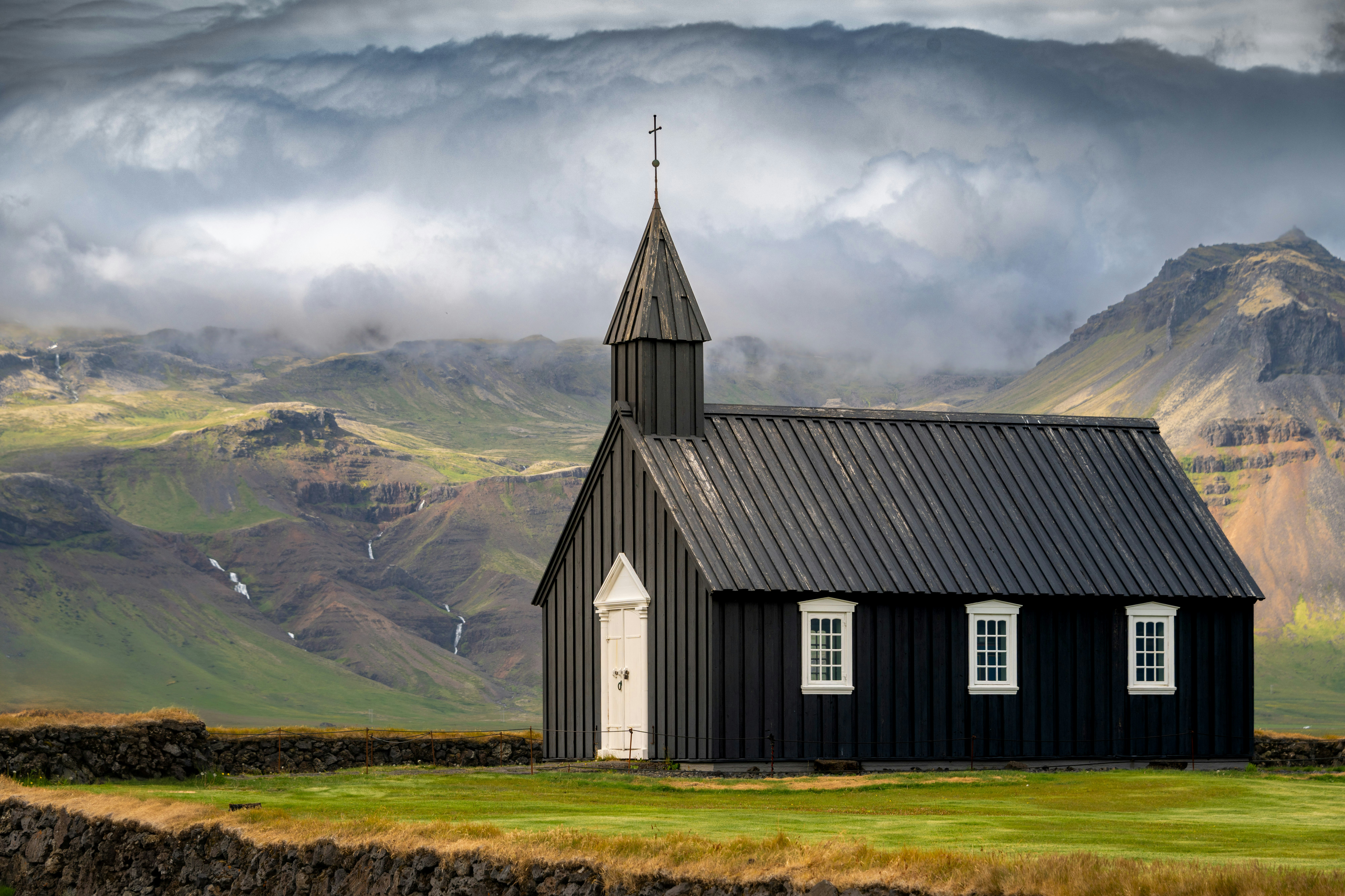 A black wooden church with white trim against a backdrop of green mountains