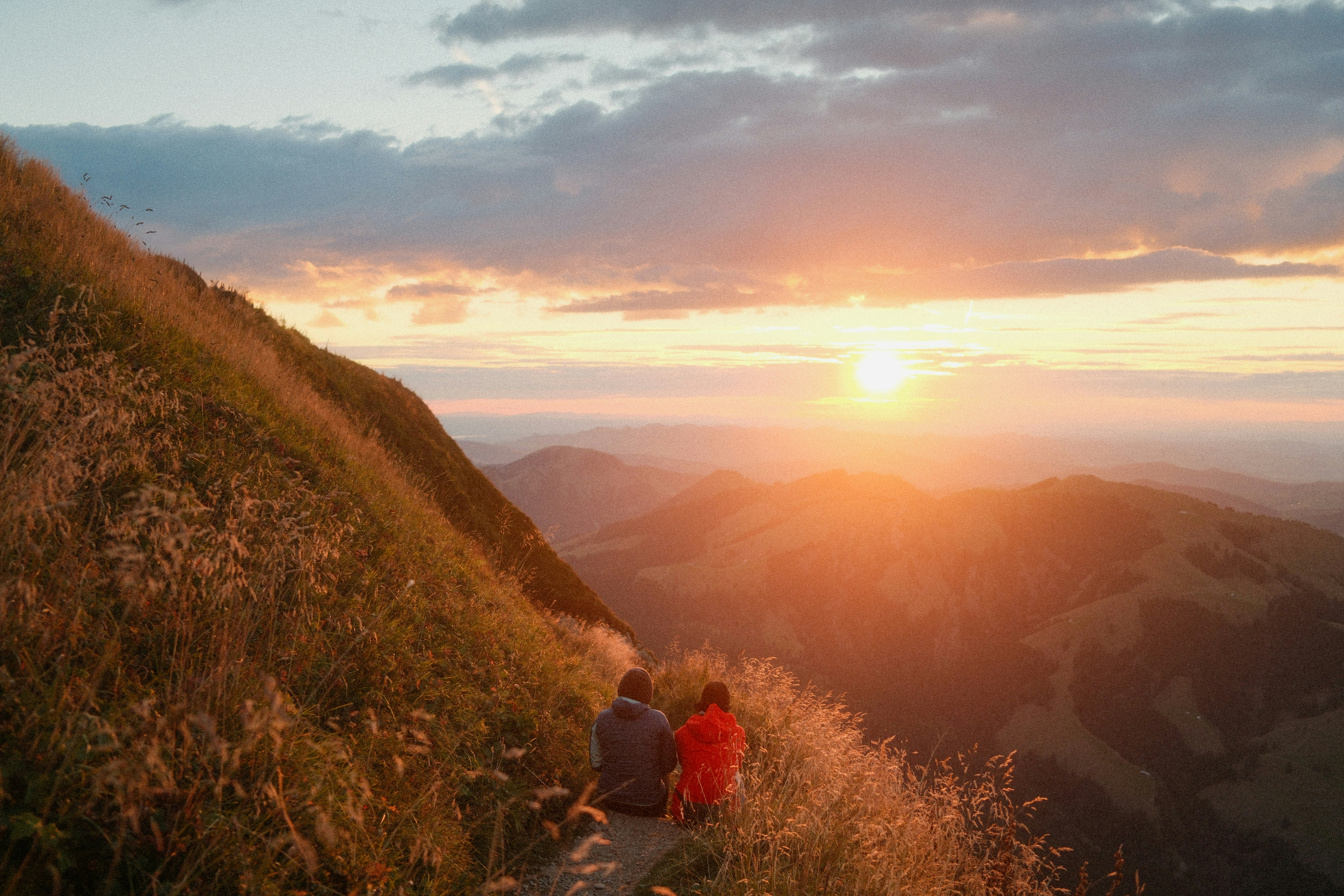 Two people sitting on a grassy mountain path in Schwende-Rüte, Switzerland, at golden hour