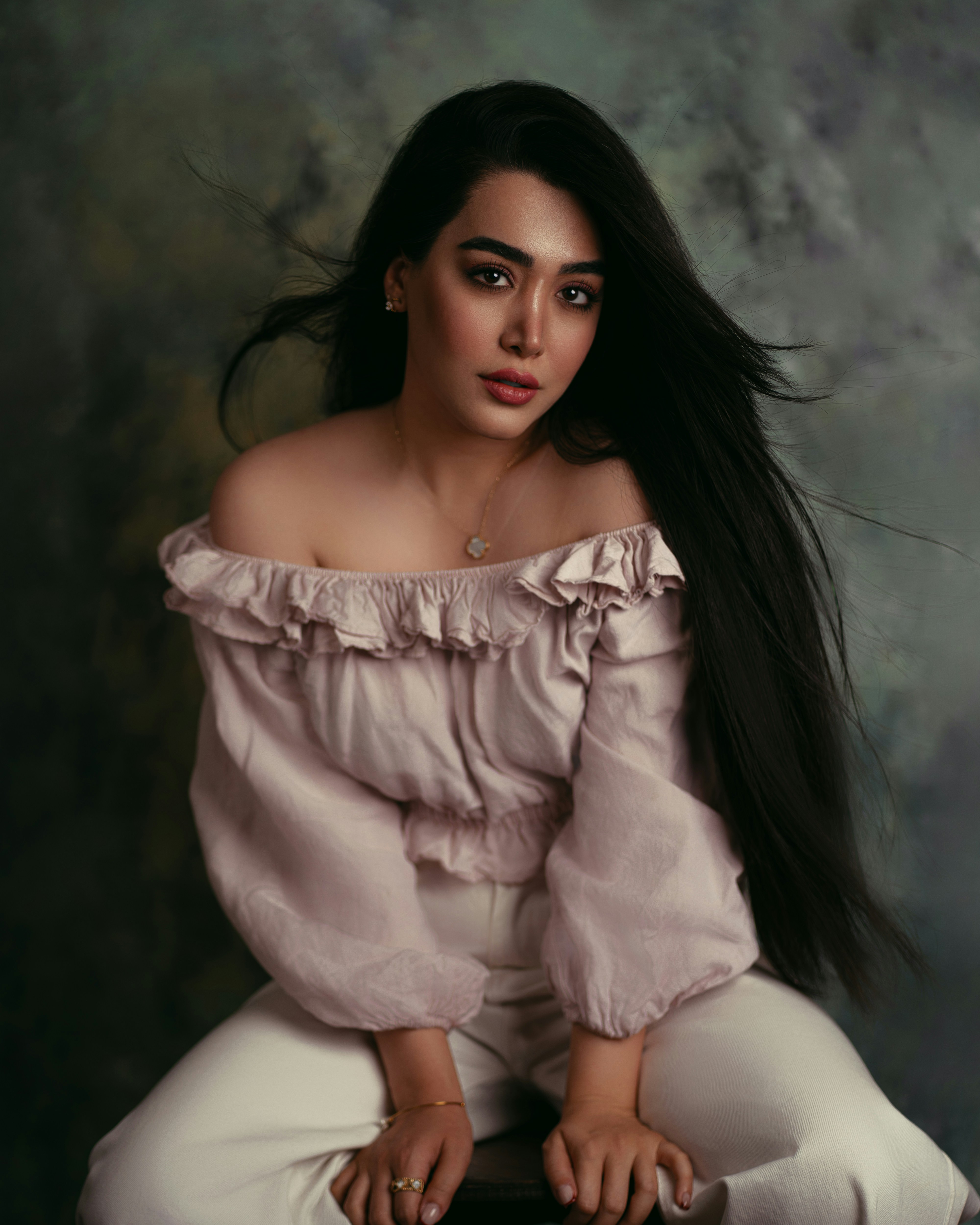 A woman with long dark hair wearing a ruffled off-shoulder top and white trousers