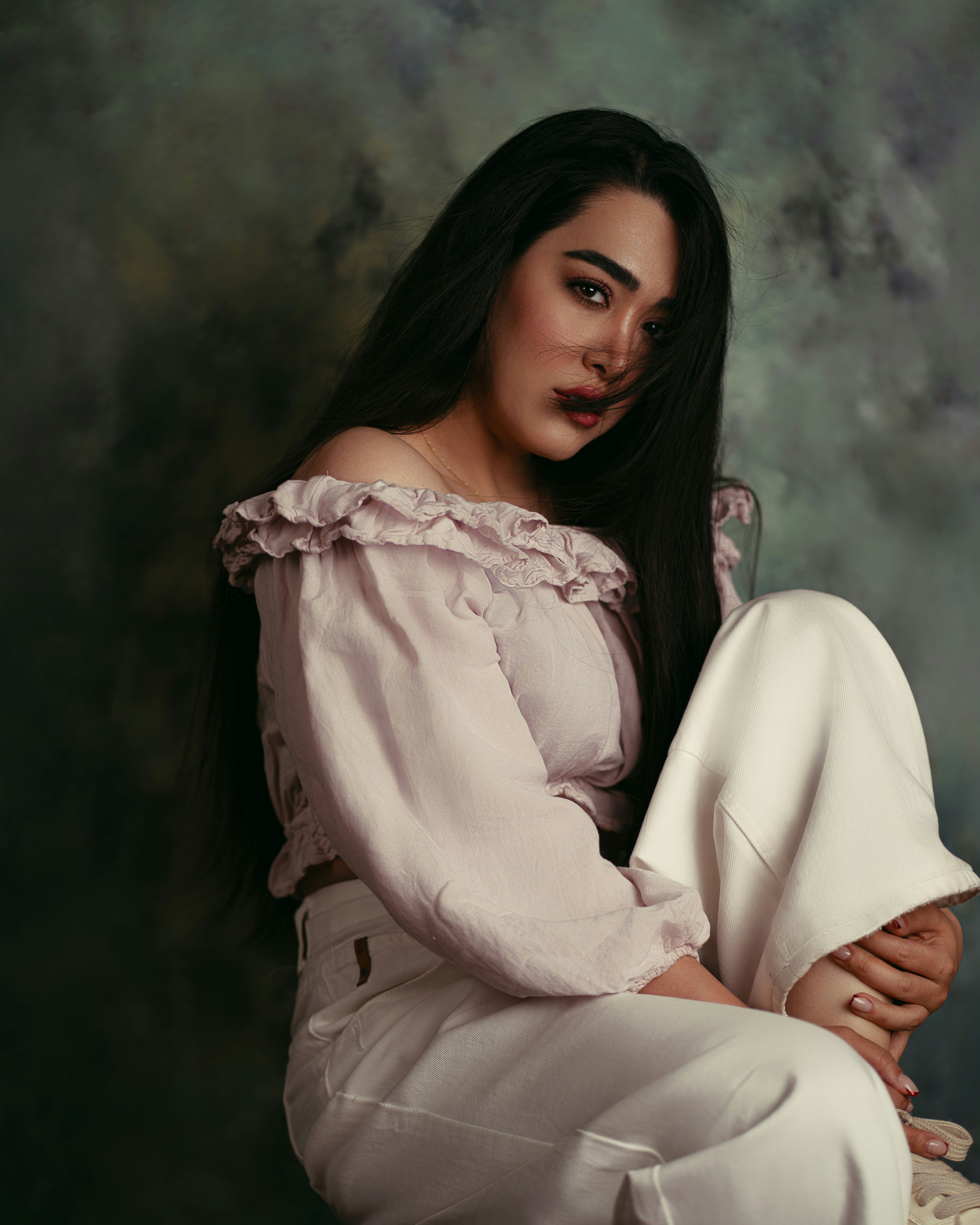 A woman with long dark hair wearing a ruffled off-the-shoulder top and white trousers