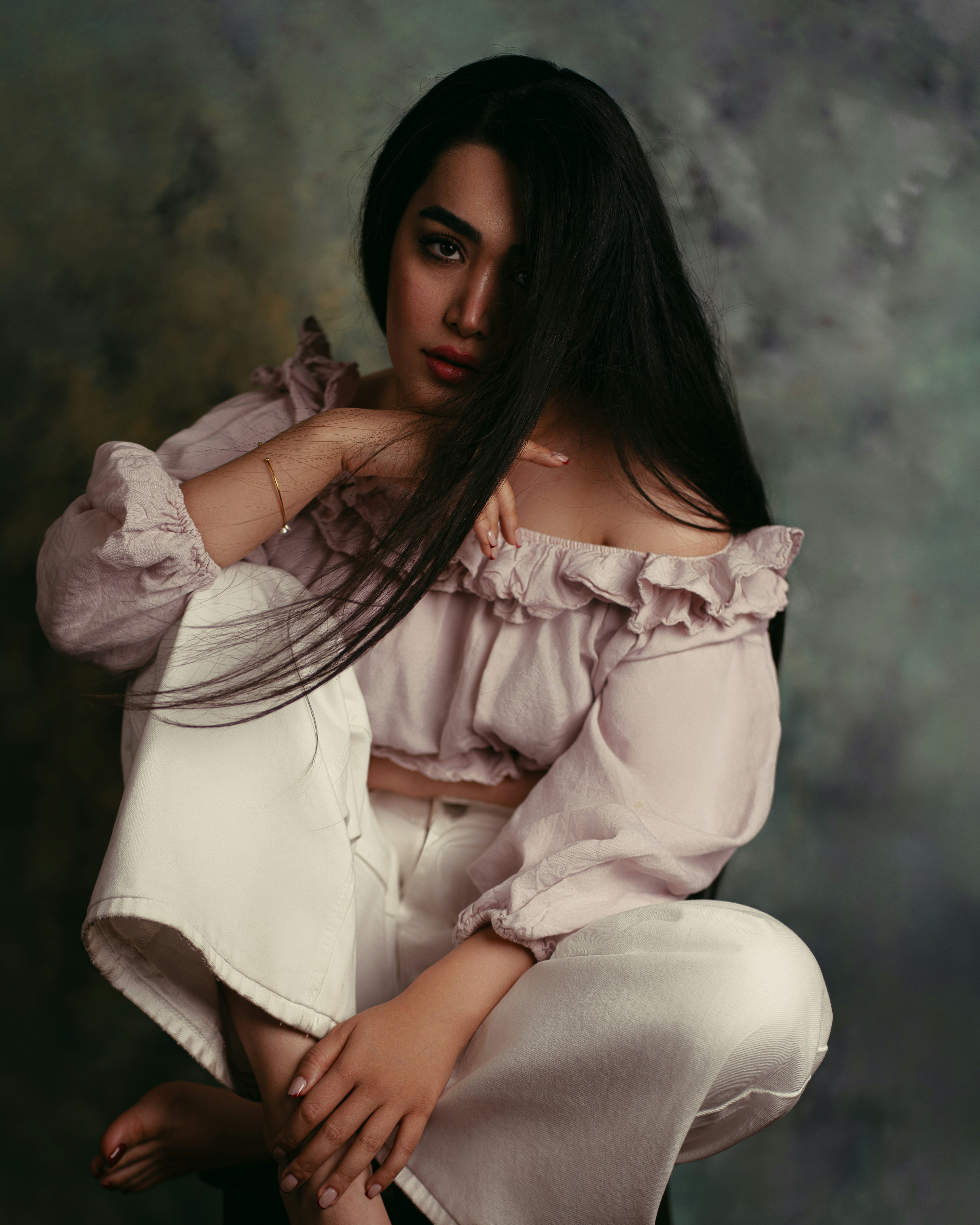 A woman with long dark hair wearing a pale pink off-shoulder top and white trousers
