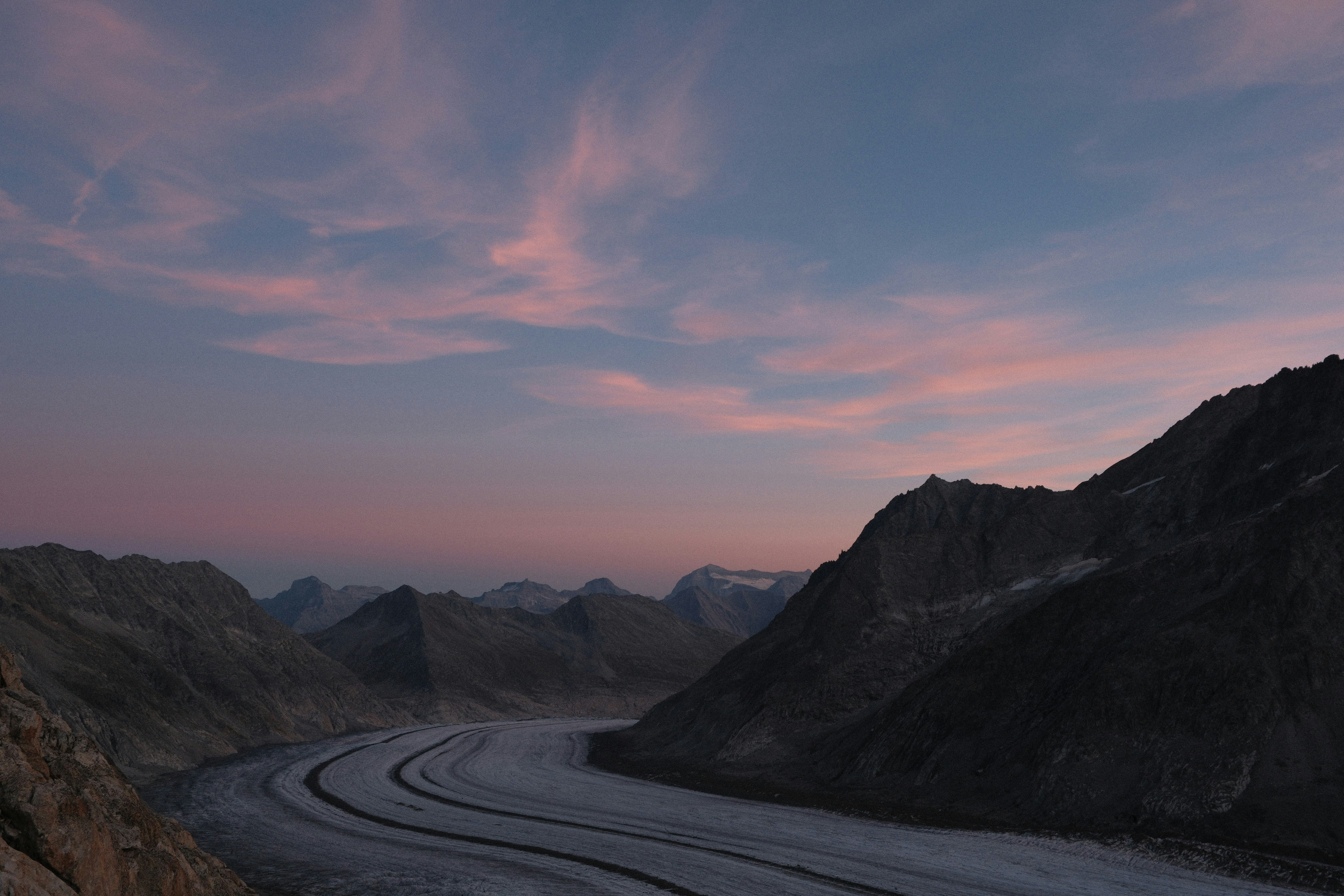 A winding glacier between dark mountain ridges under a soft pink and blue sky