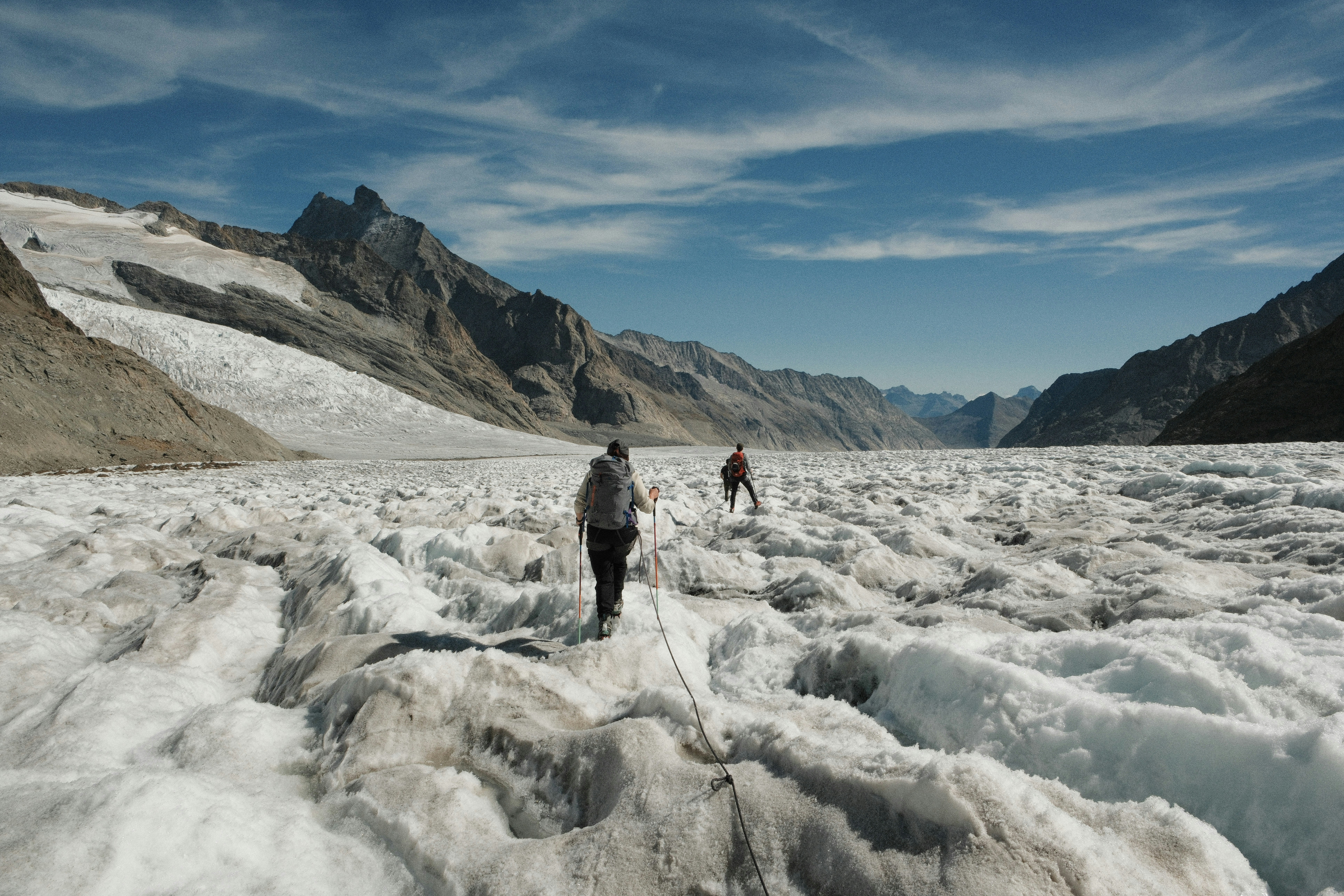 Two people hiking across a vast glacier surrounded by high rocky mountain peaks