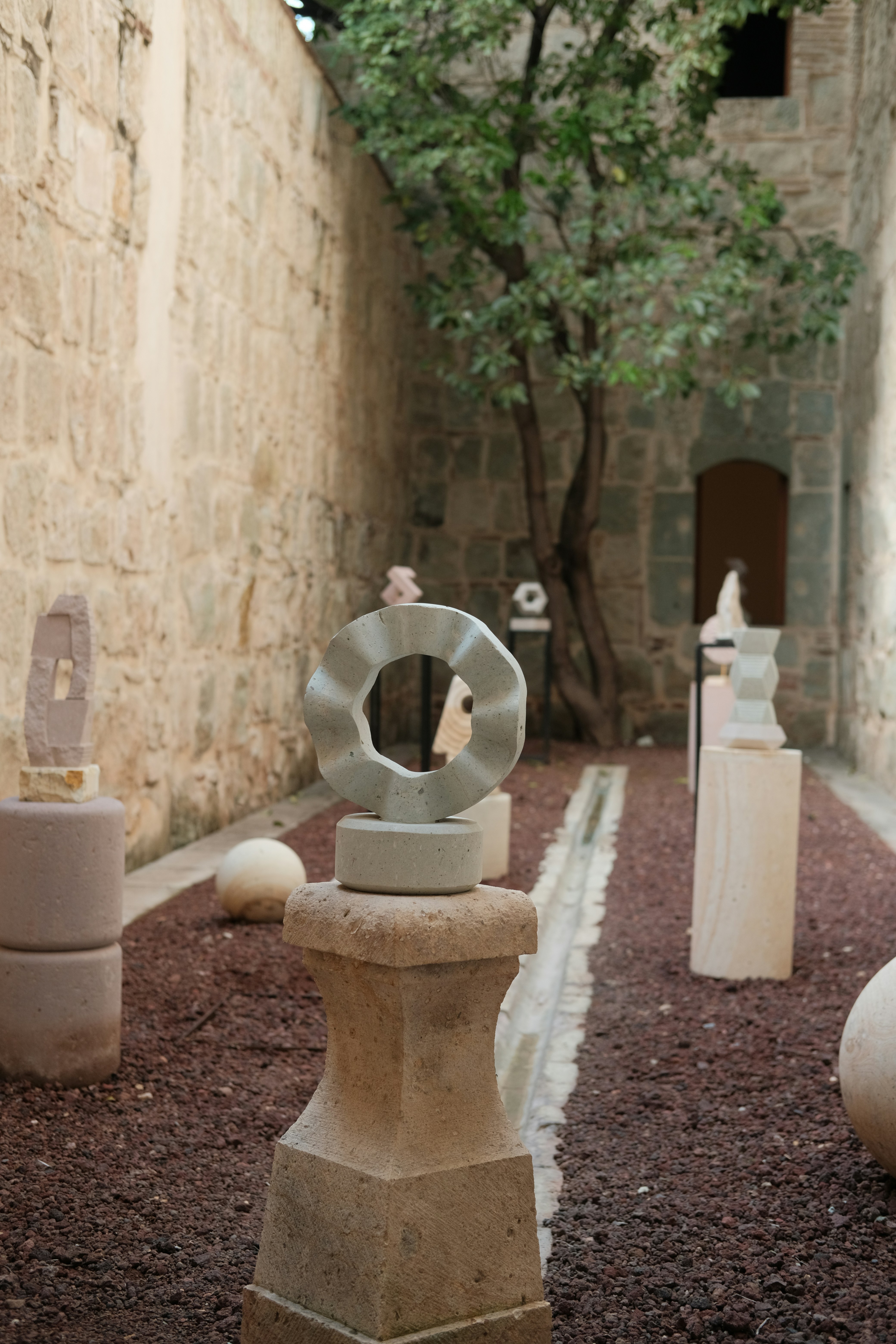 Esculturas de piedra sobre pedestales en un patio con árbol y grava