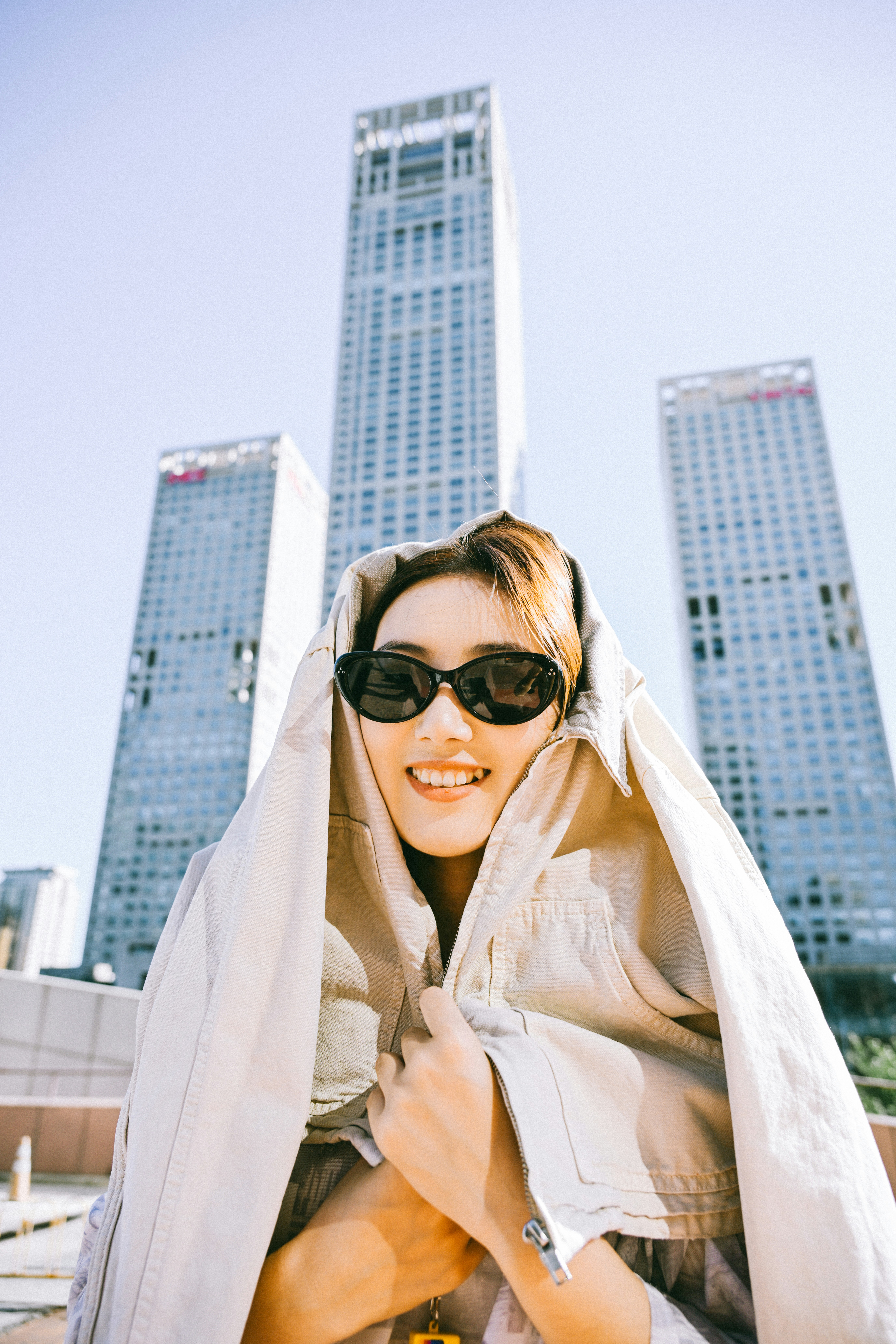 A smiling person wearing sunglasses and a beige jacket over their head in a city