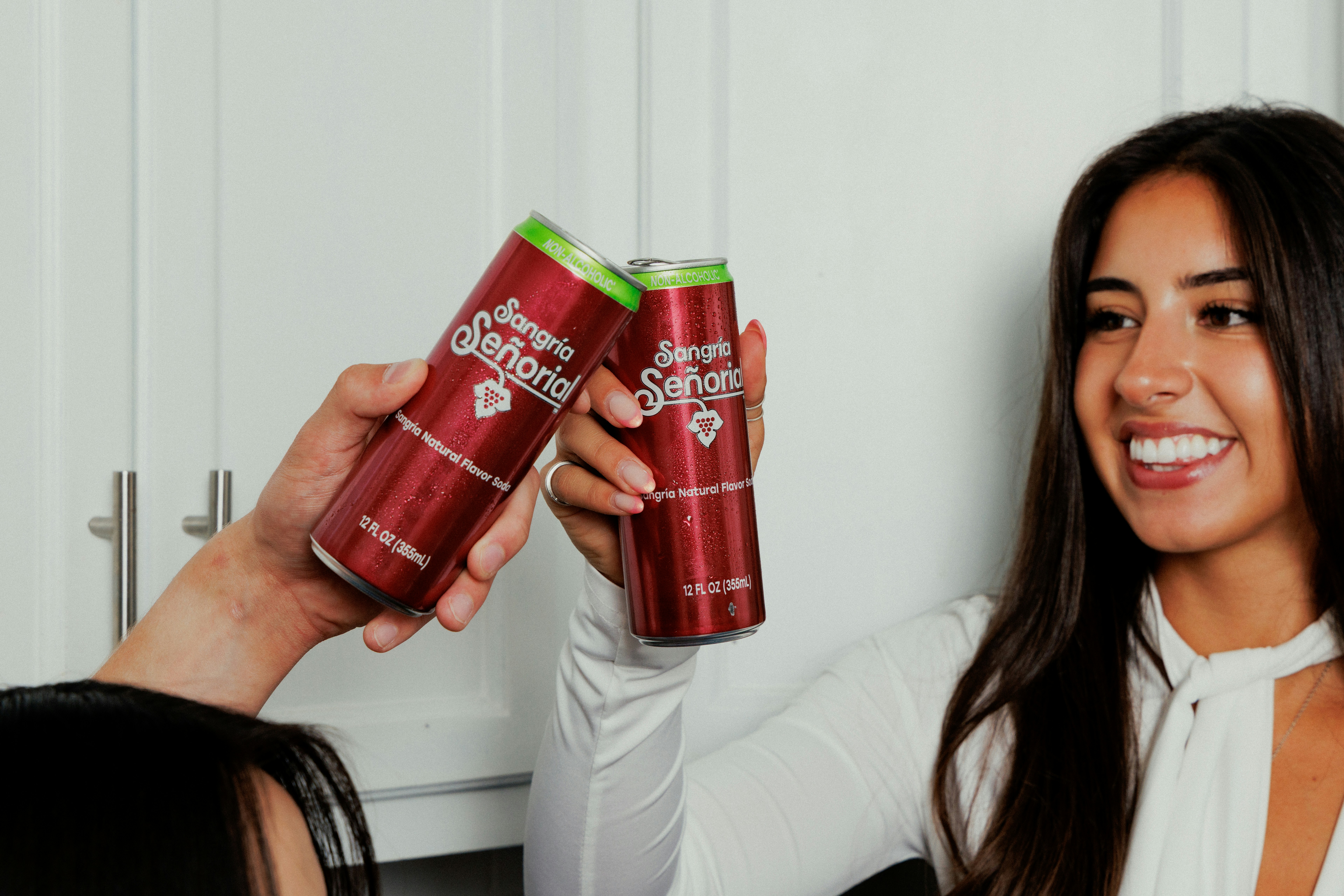 Two people toasting with cans of Sangria Señorial soda in a bright kitchen