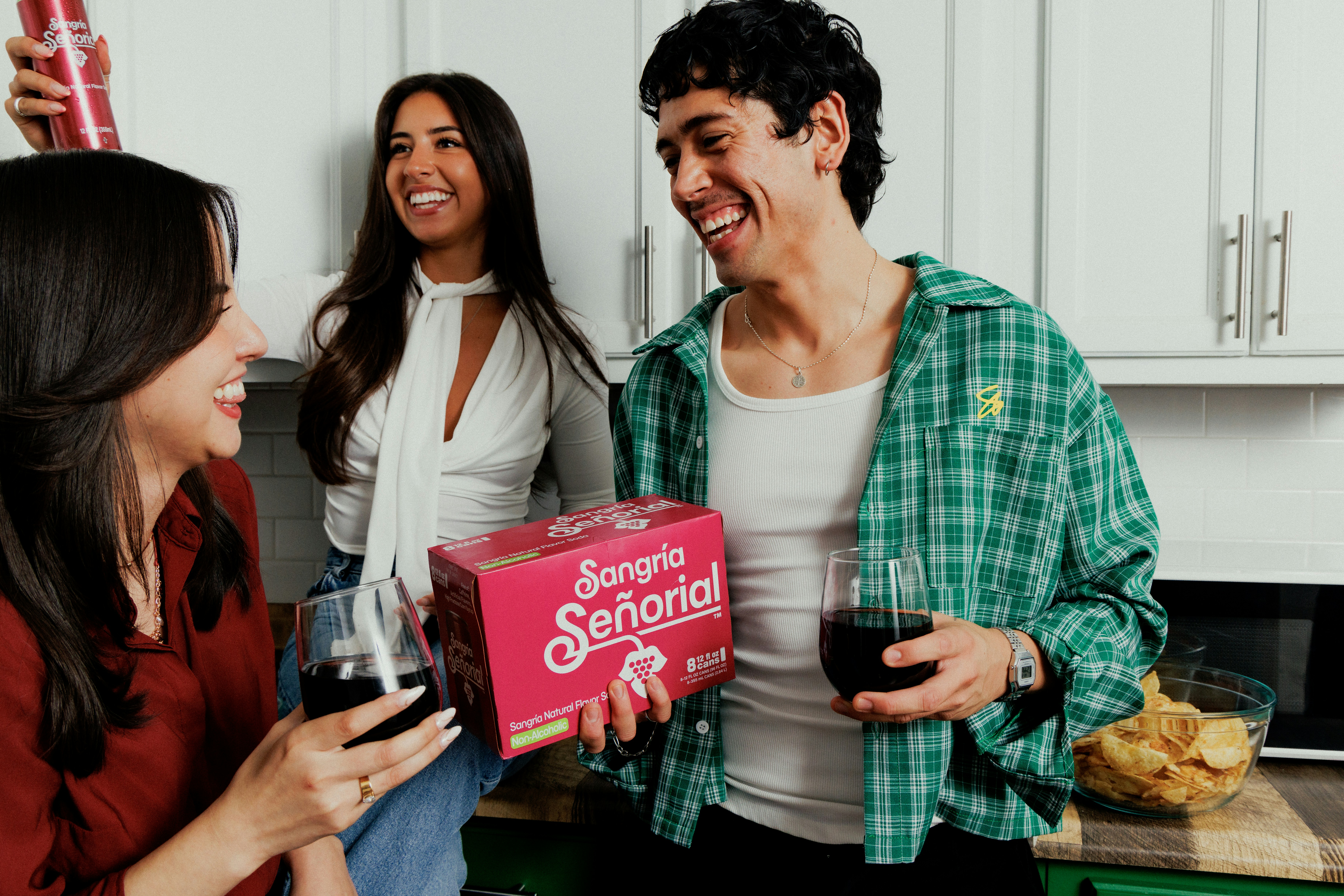 Three people laughing while holding glasses of red soda in a bright kitchen