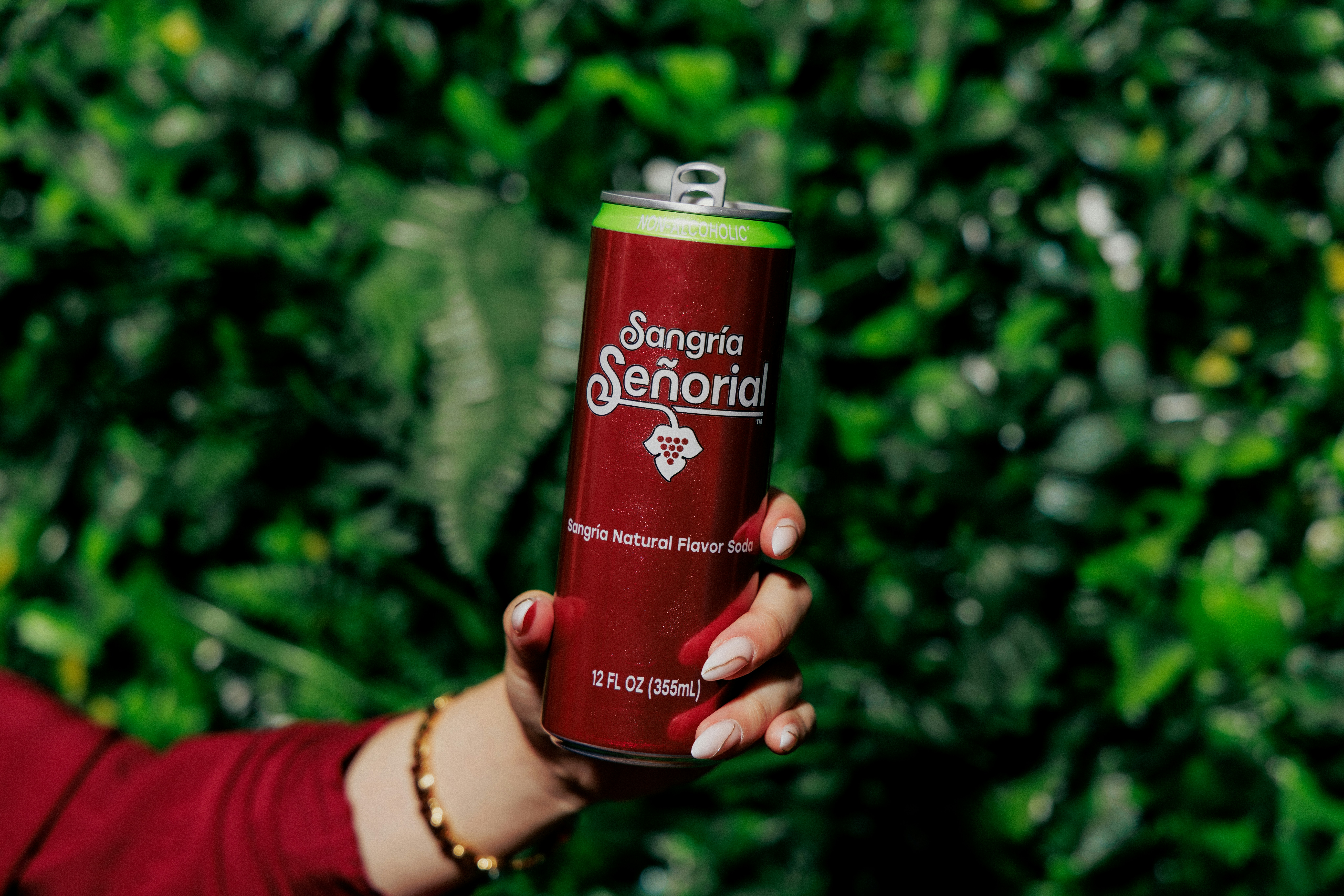 A hand holding a red can of Sangria Señorial soda against green foliage
