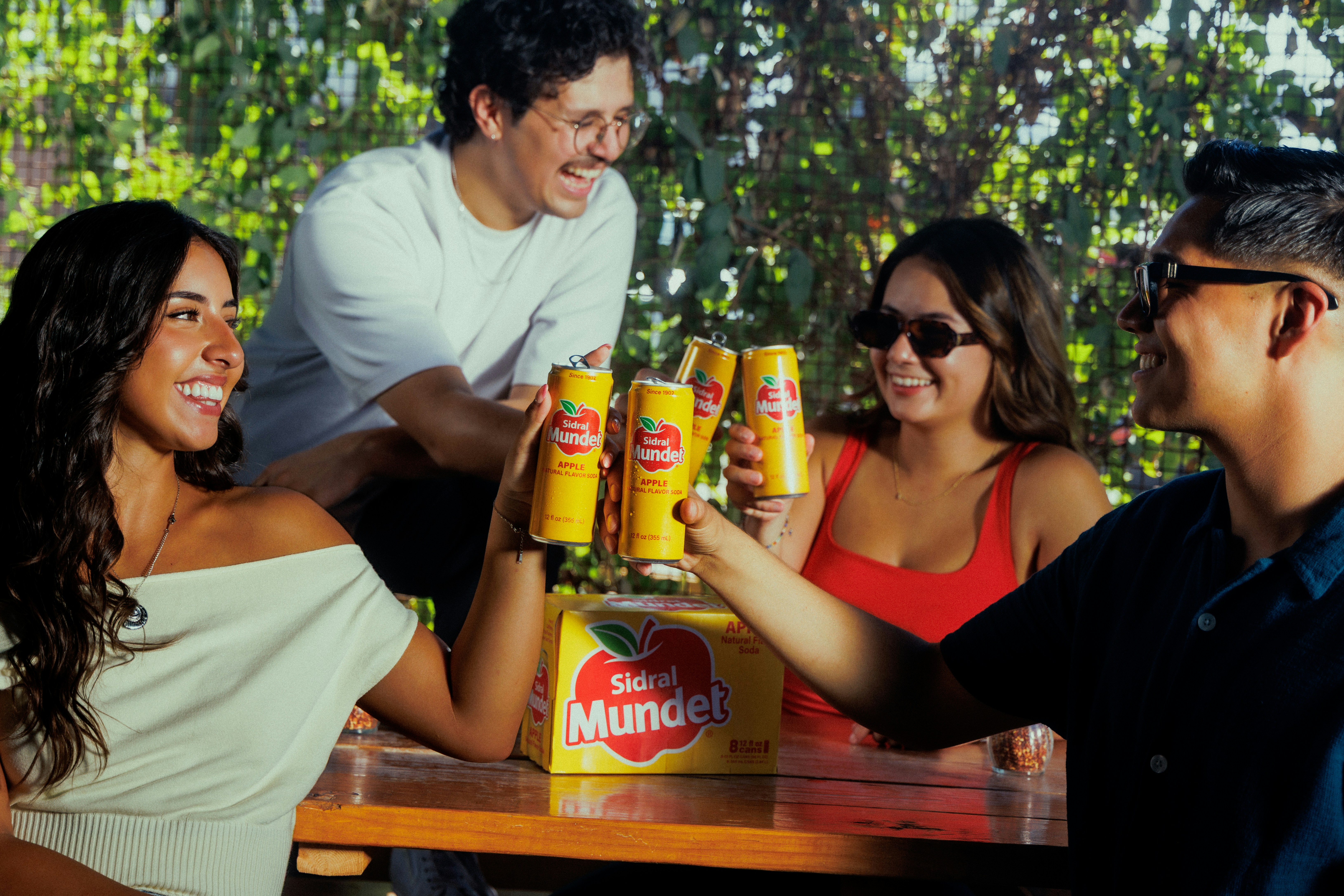 Four people toasting with yellow cans of Sidral Mundet soda at a wooden table