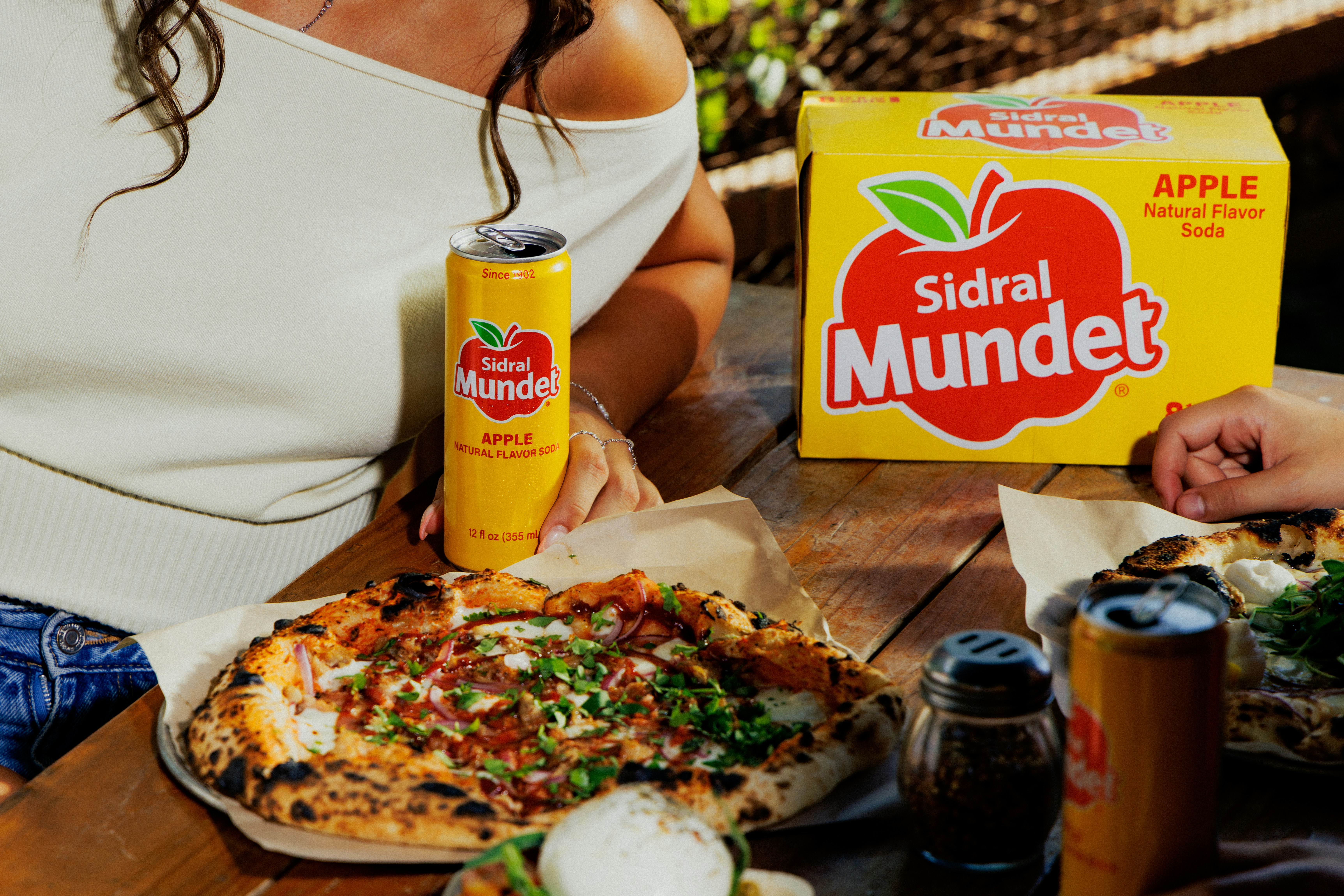 A person holding a can of Sidral Mundet soda beside pizza on a wooden table