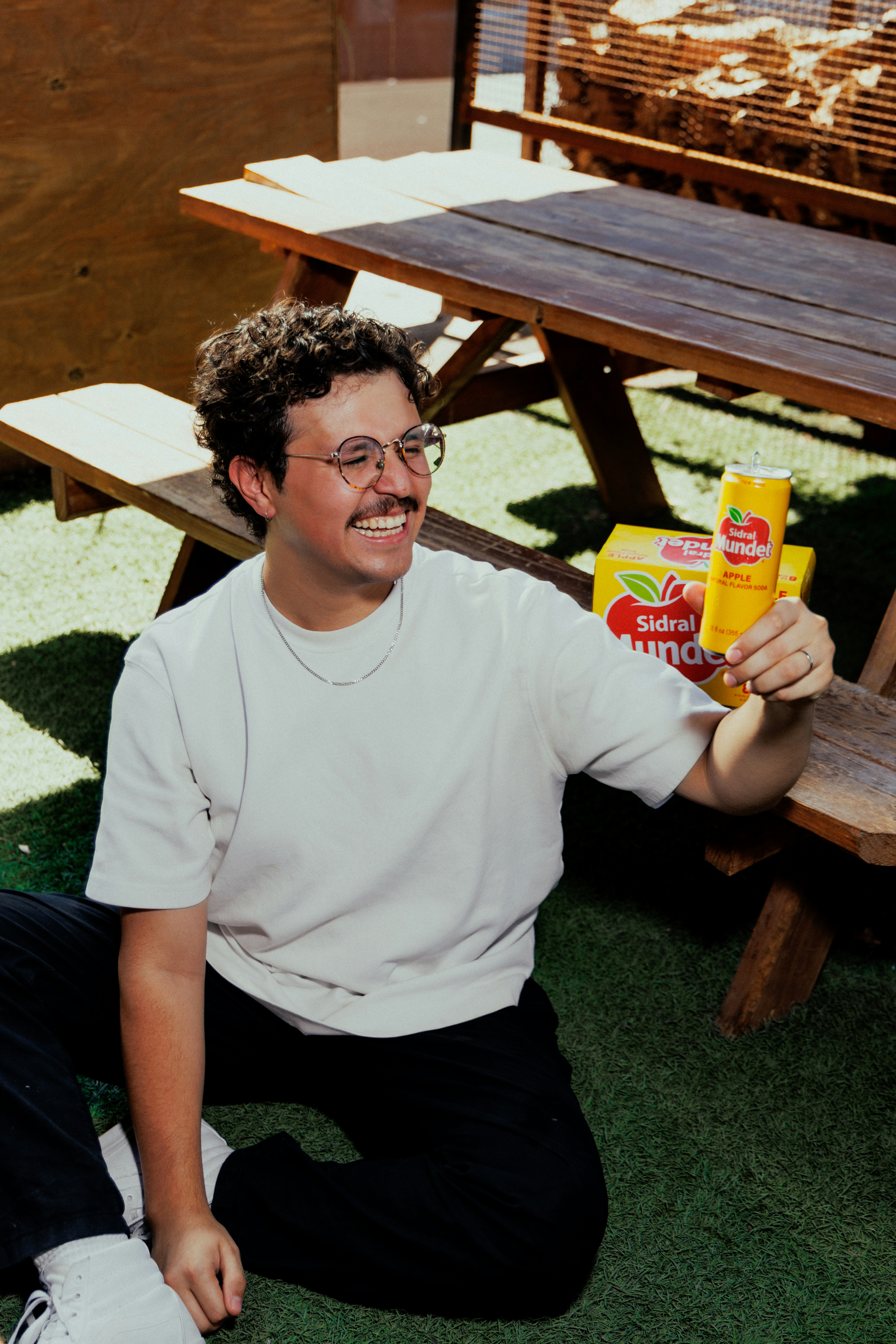 A smiling person in a white t-shirt holding a yellow soda can