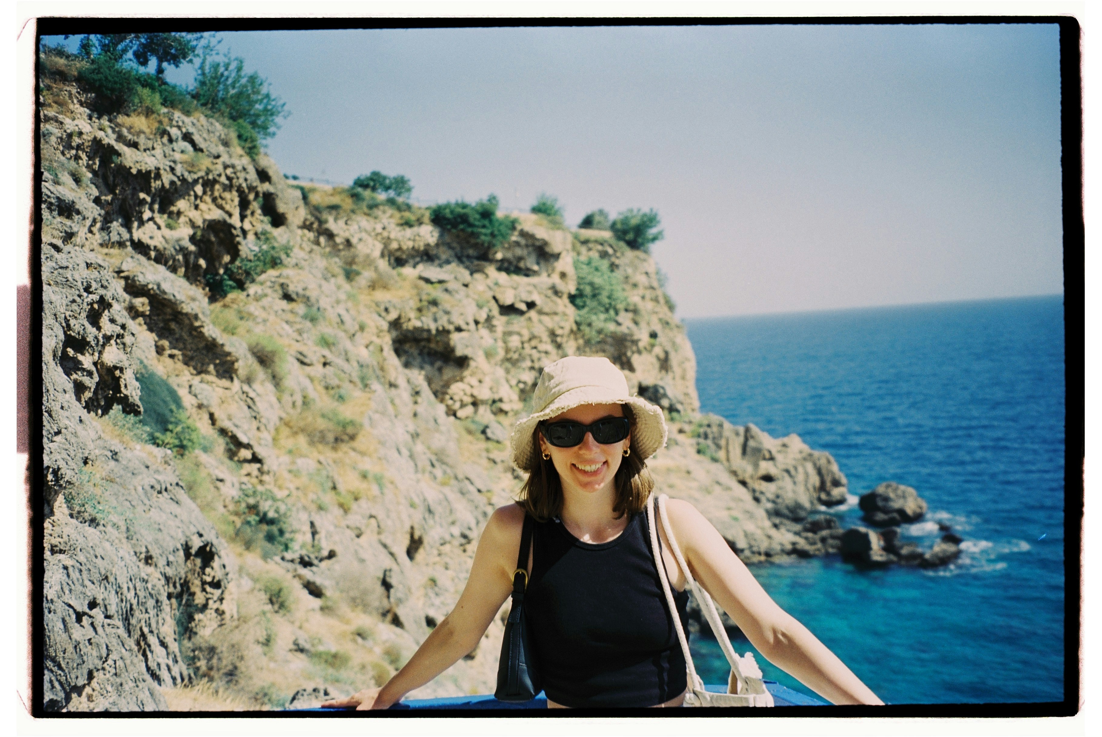 A woman in a black tank top and sun hat against a rocky coastal cliff