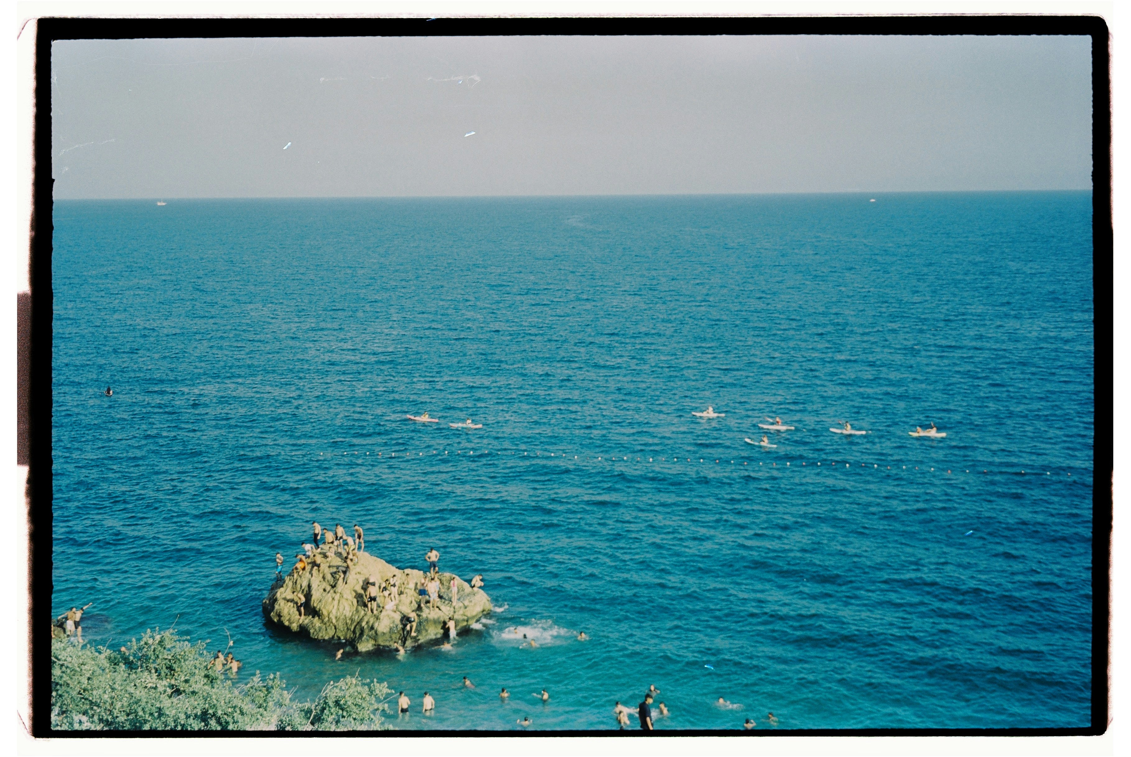 People on a rocky outcrop in the sea with others swimming and kayaking nearby