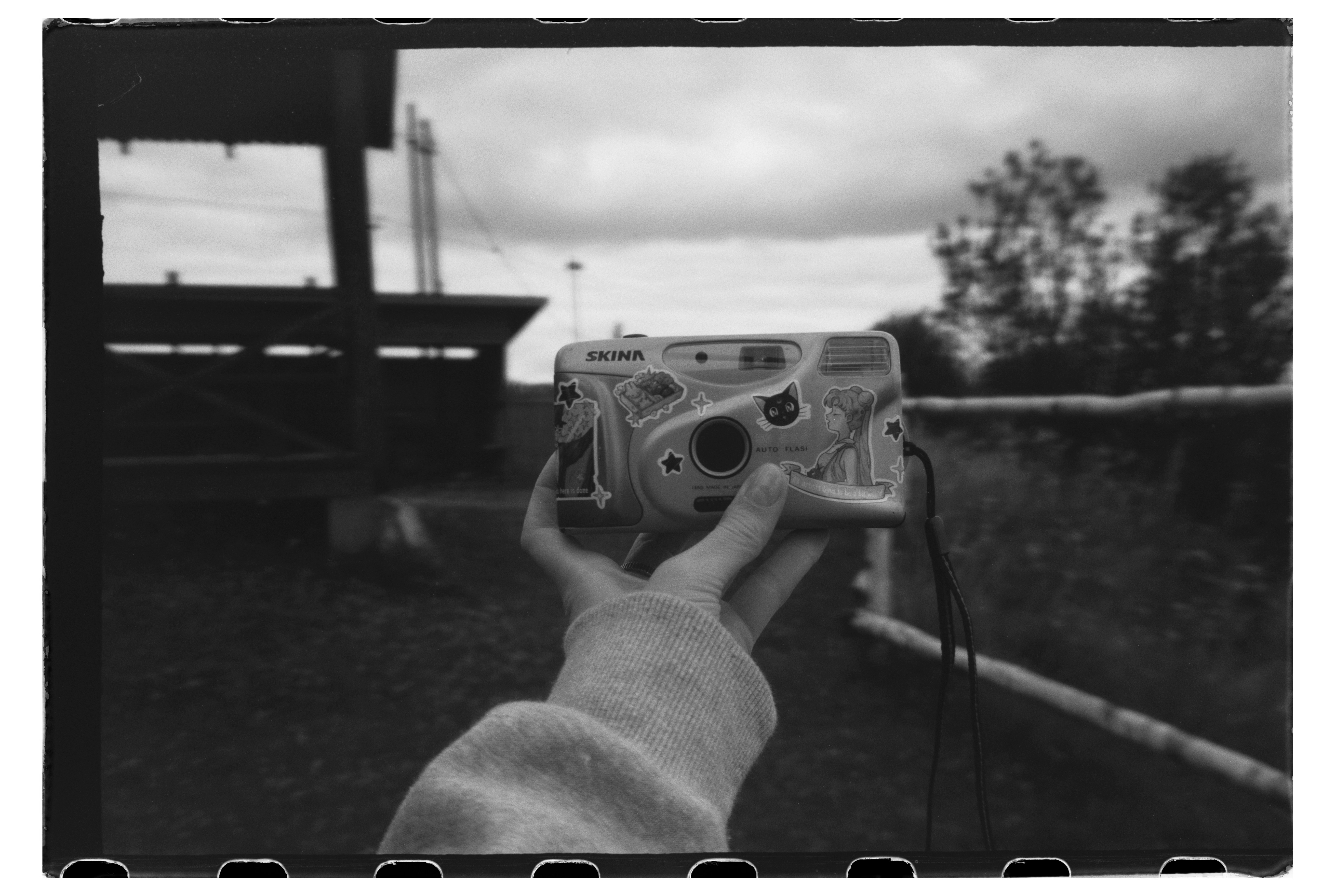 A hand holding a sticker-covered film camera against a blurred outdoor landscape