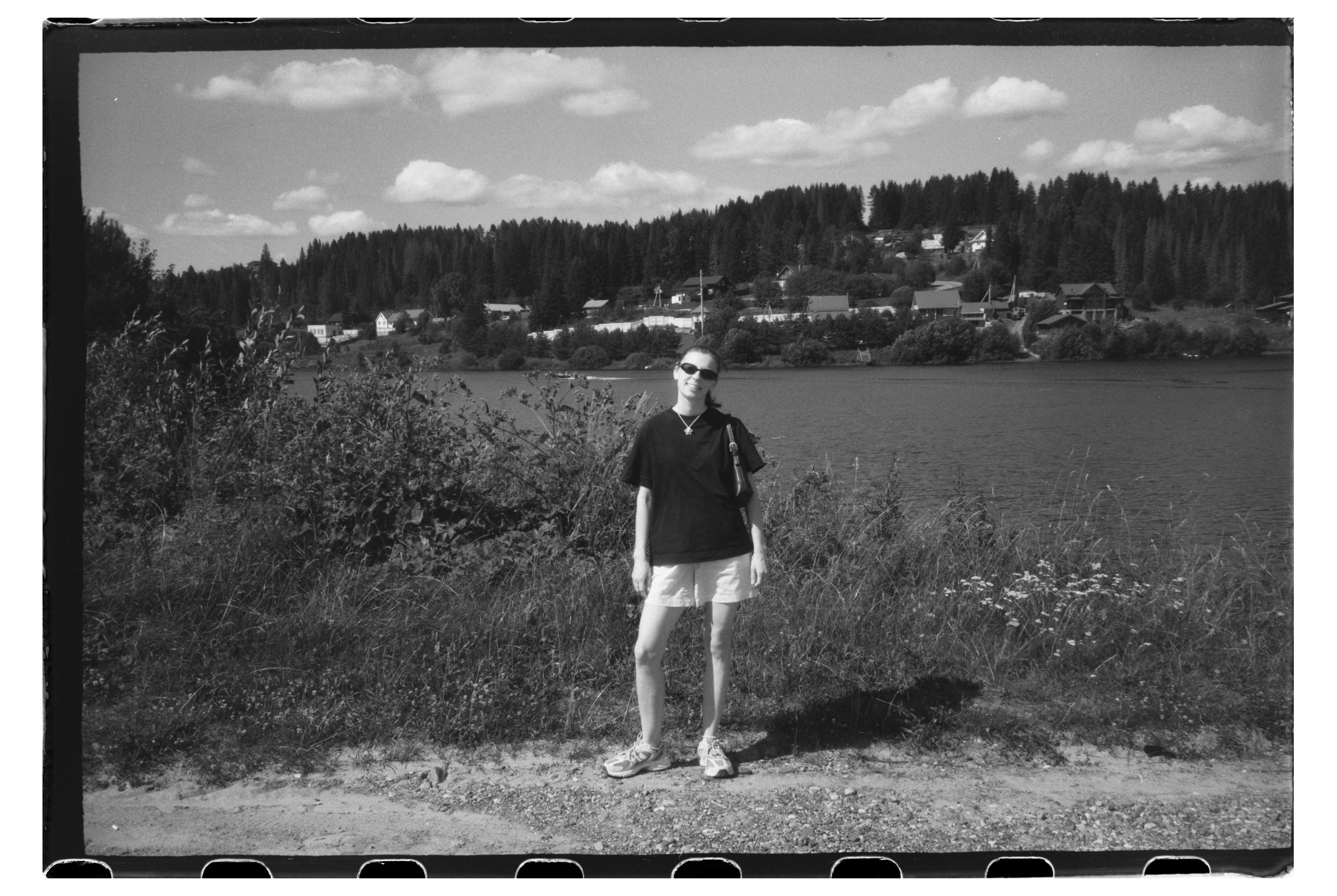A woman in sunglasses and a t-shirt standing by a lake with forest