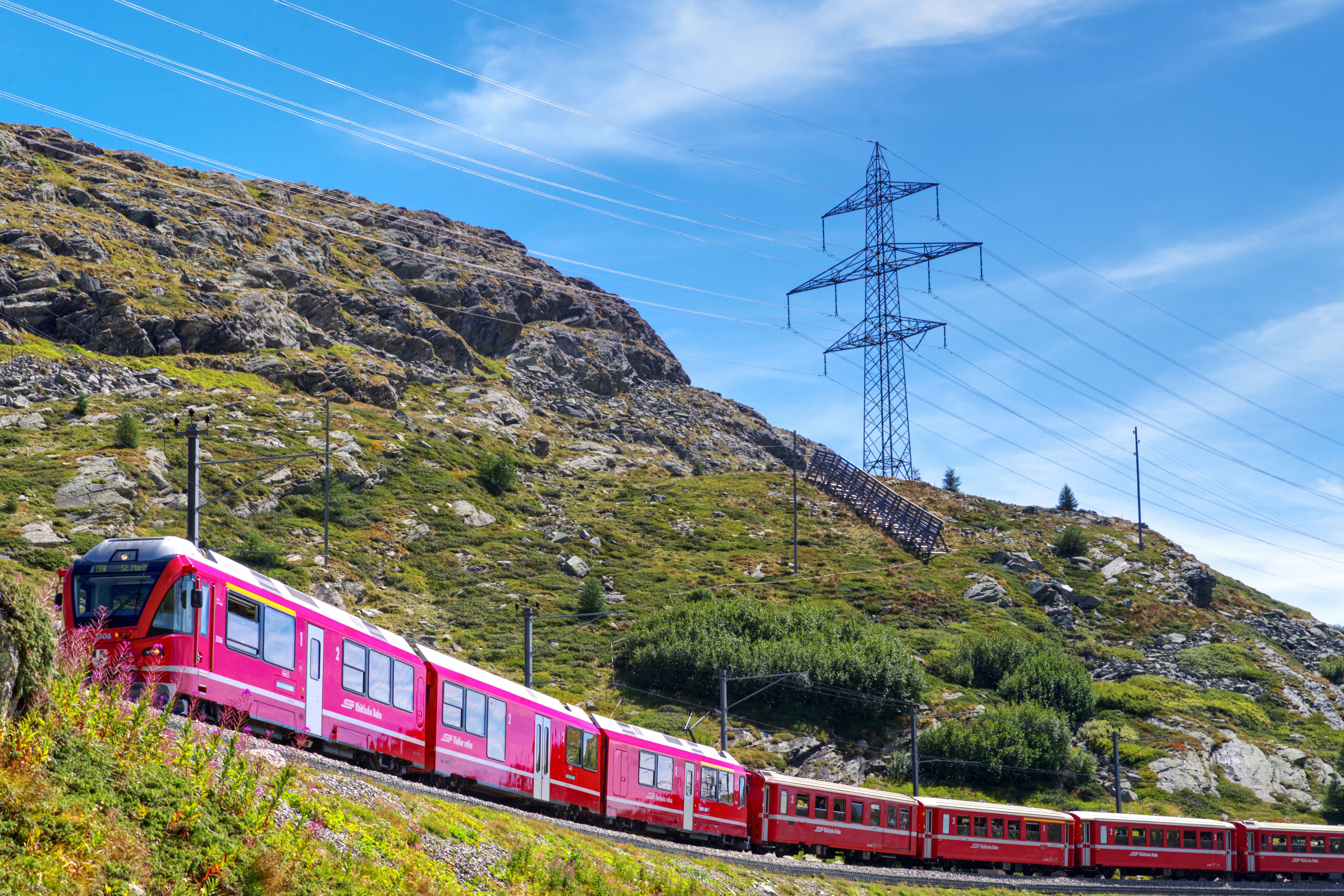 A red train traveling along a mountain track beneath a large electrical pylon