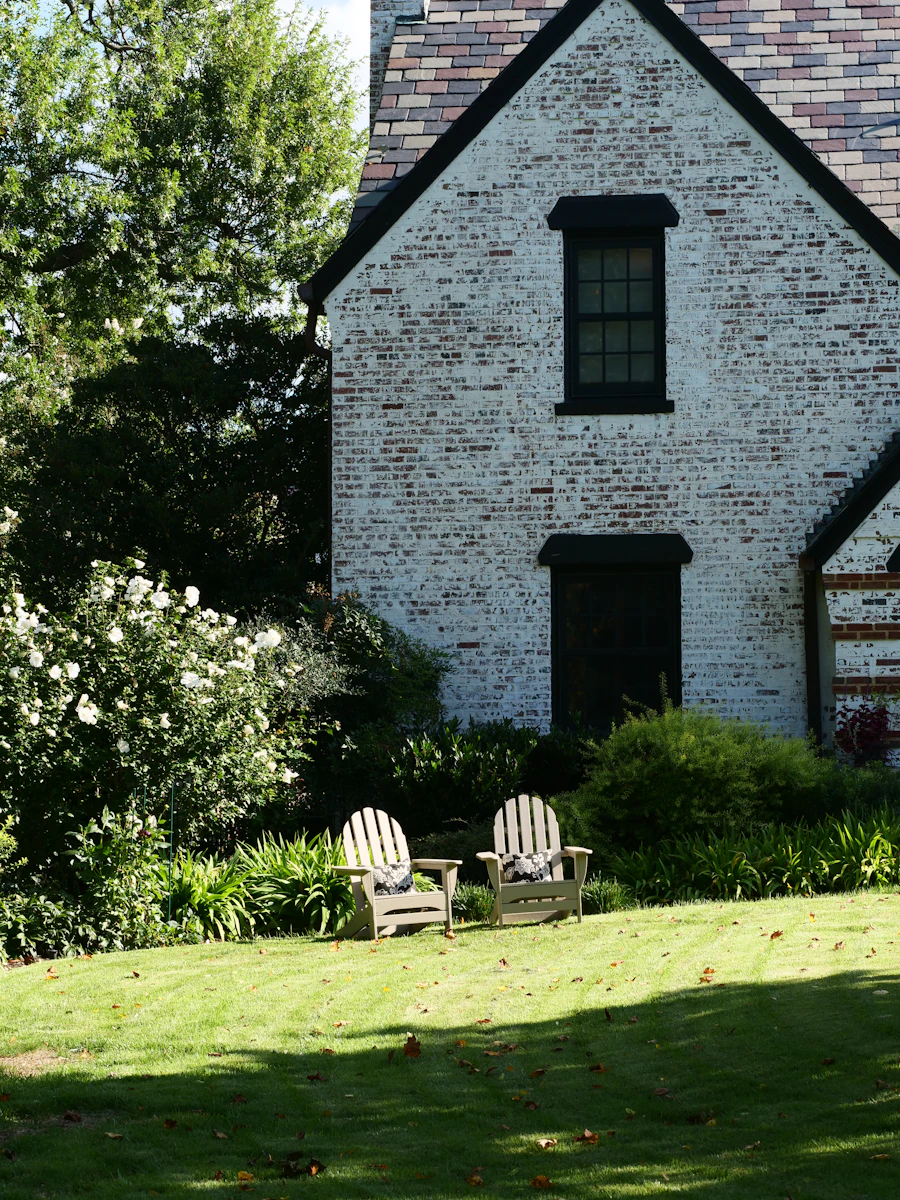 Mature residential garden with a green lawn, two white garden chairs and flowering borders in front of a stone house