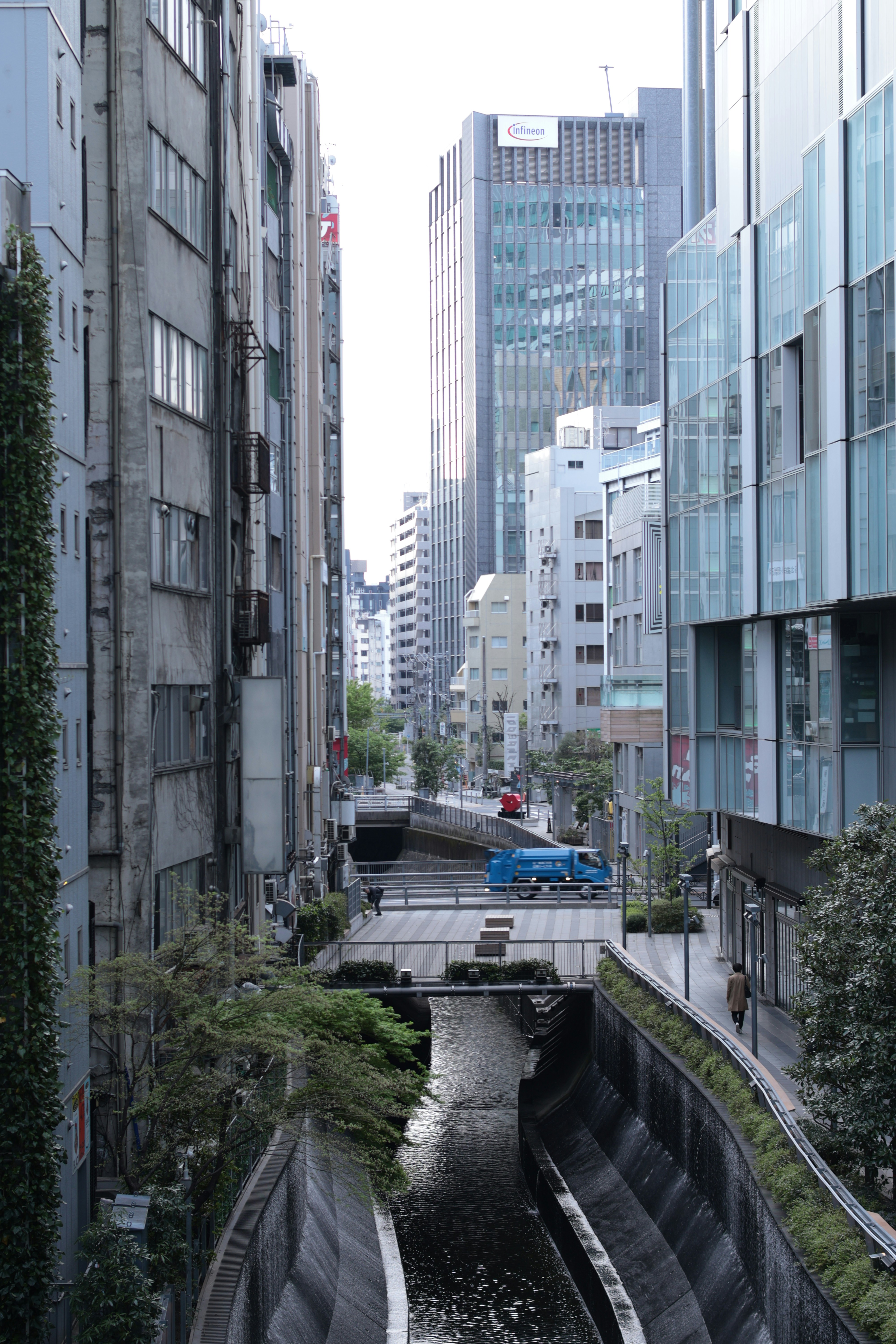 A narrow canal flowing between tall urban buildings with a blue truck crossing a bridge