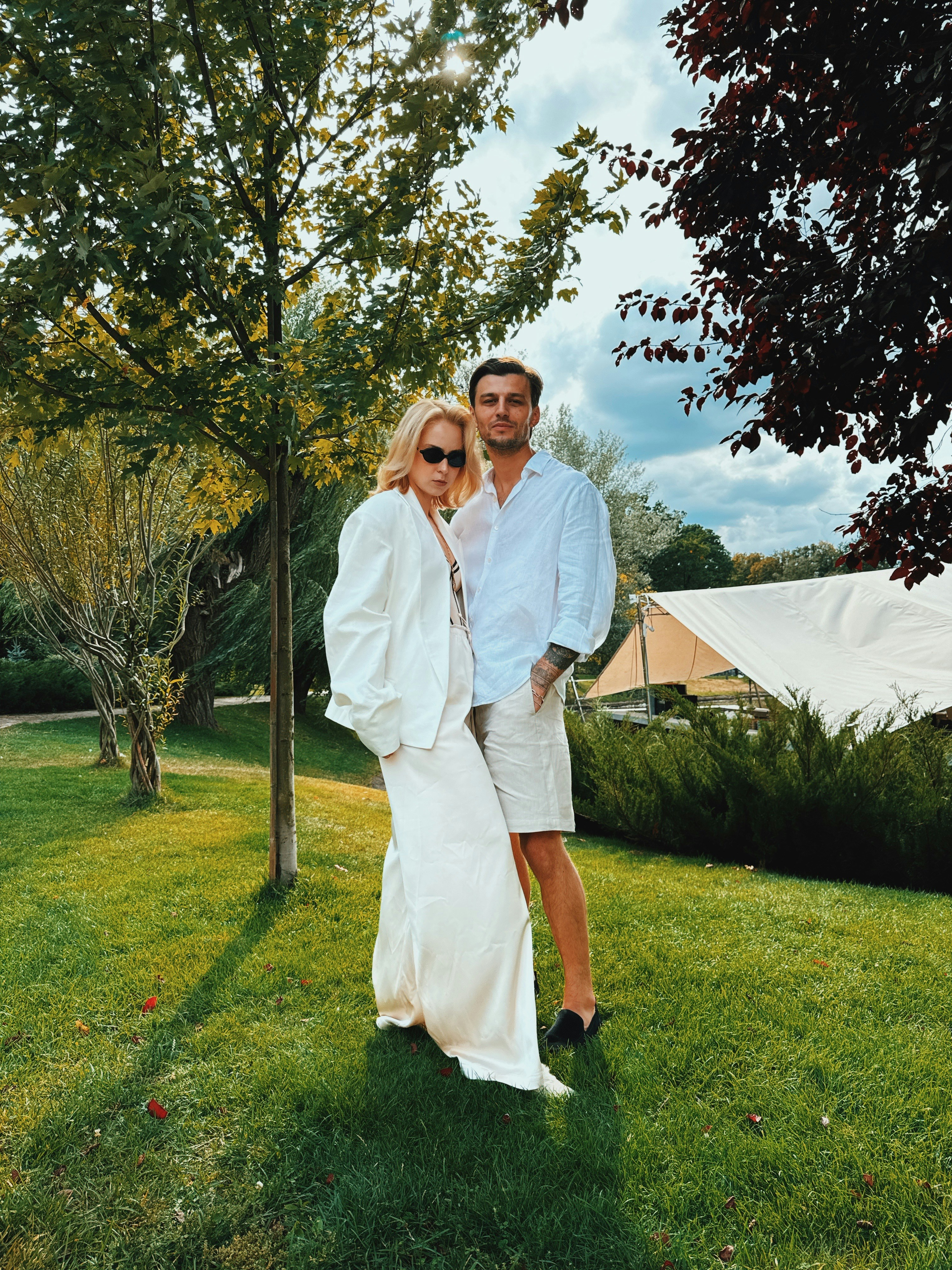 A man and a woman in white clothing standing on green grass under trees