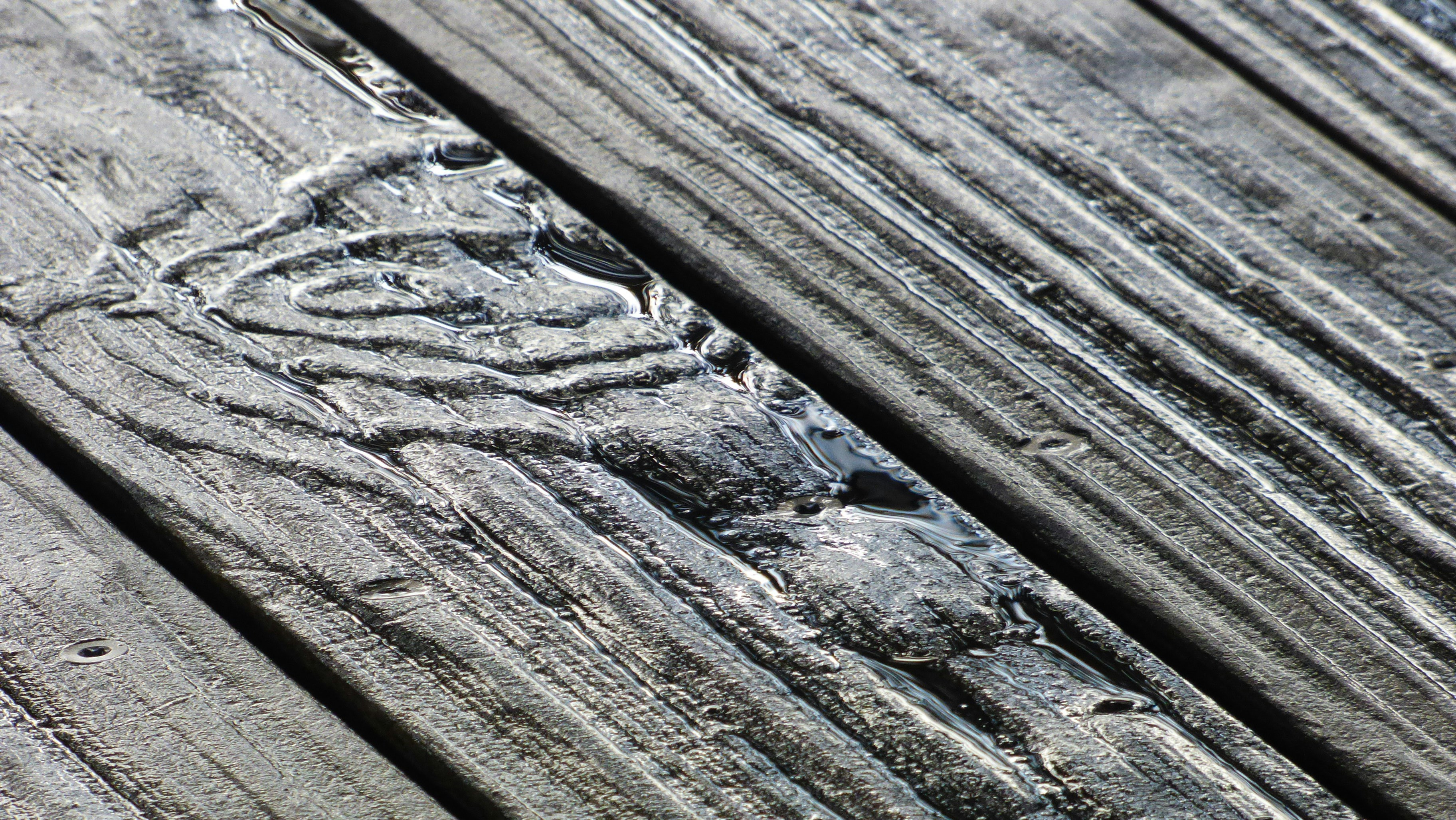 Weathered gray wooden deck boards with rainwater pooling in the grain, showing sun and moisture damage before staining