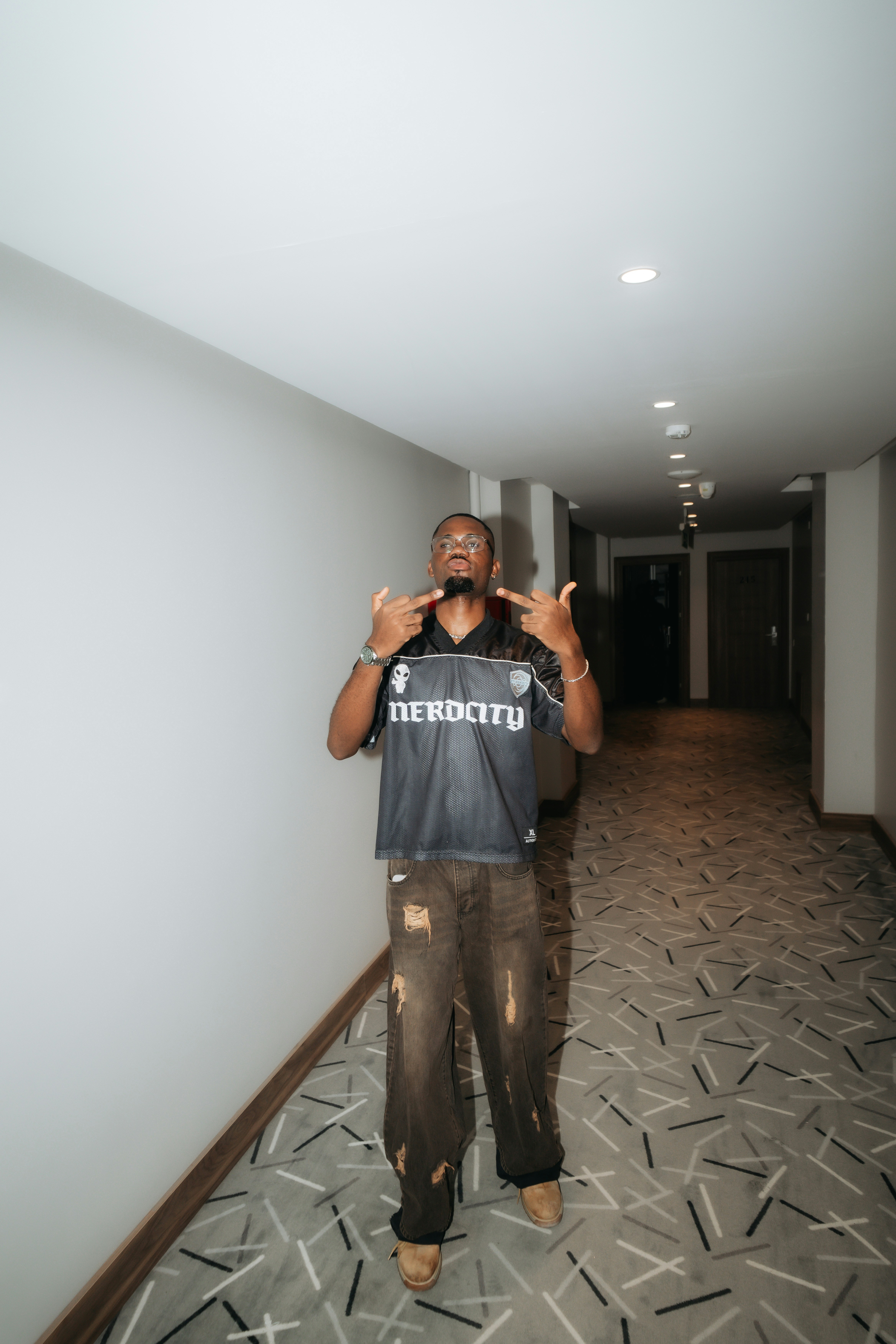 A man in a black jersey and distressed brown jeans in a hotel hallway