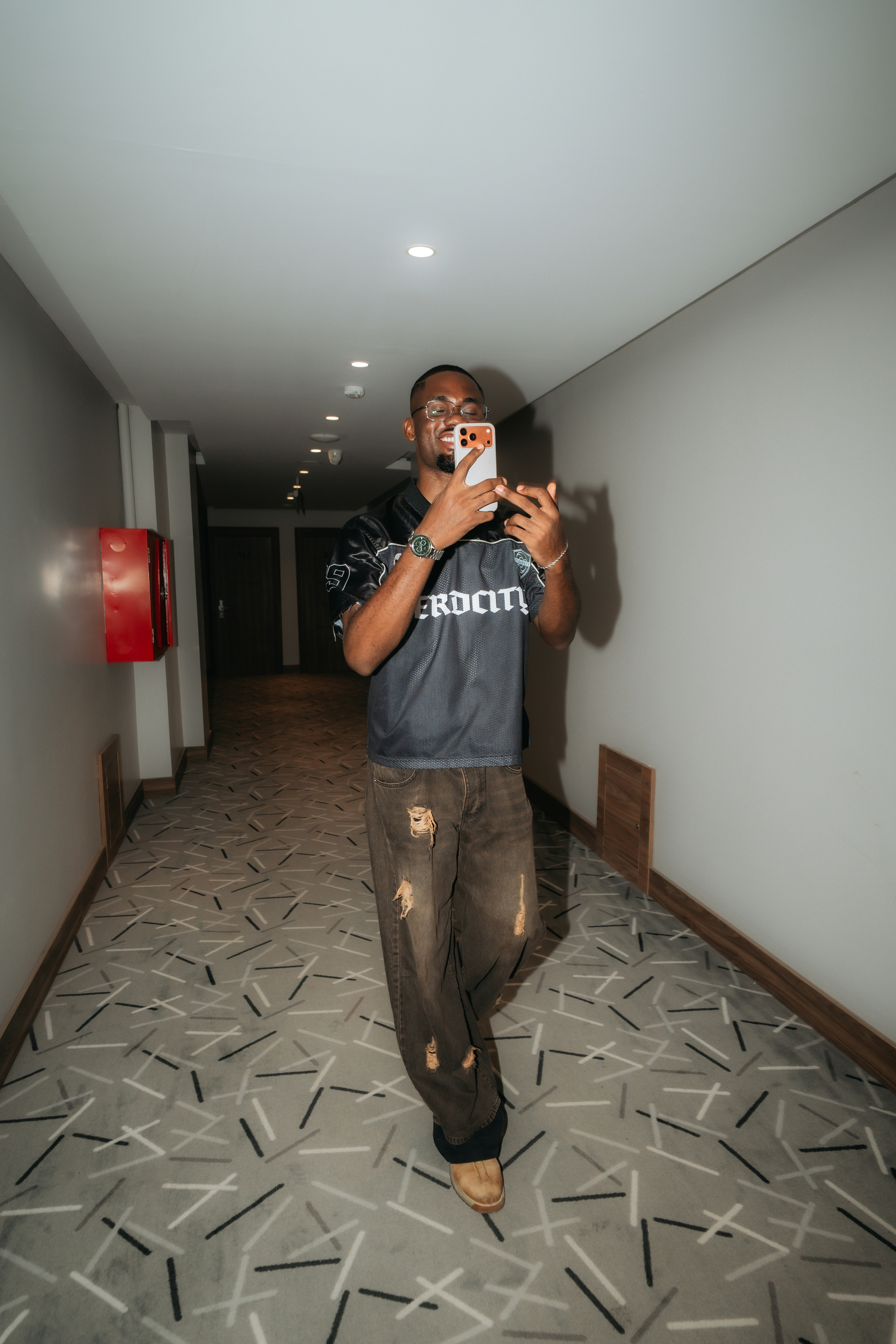 A man in a black jersey and distressed jeans taking a selfie in a hallway