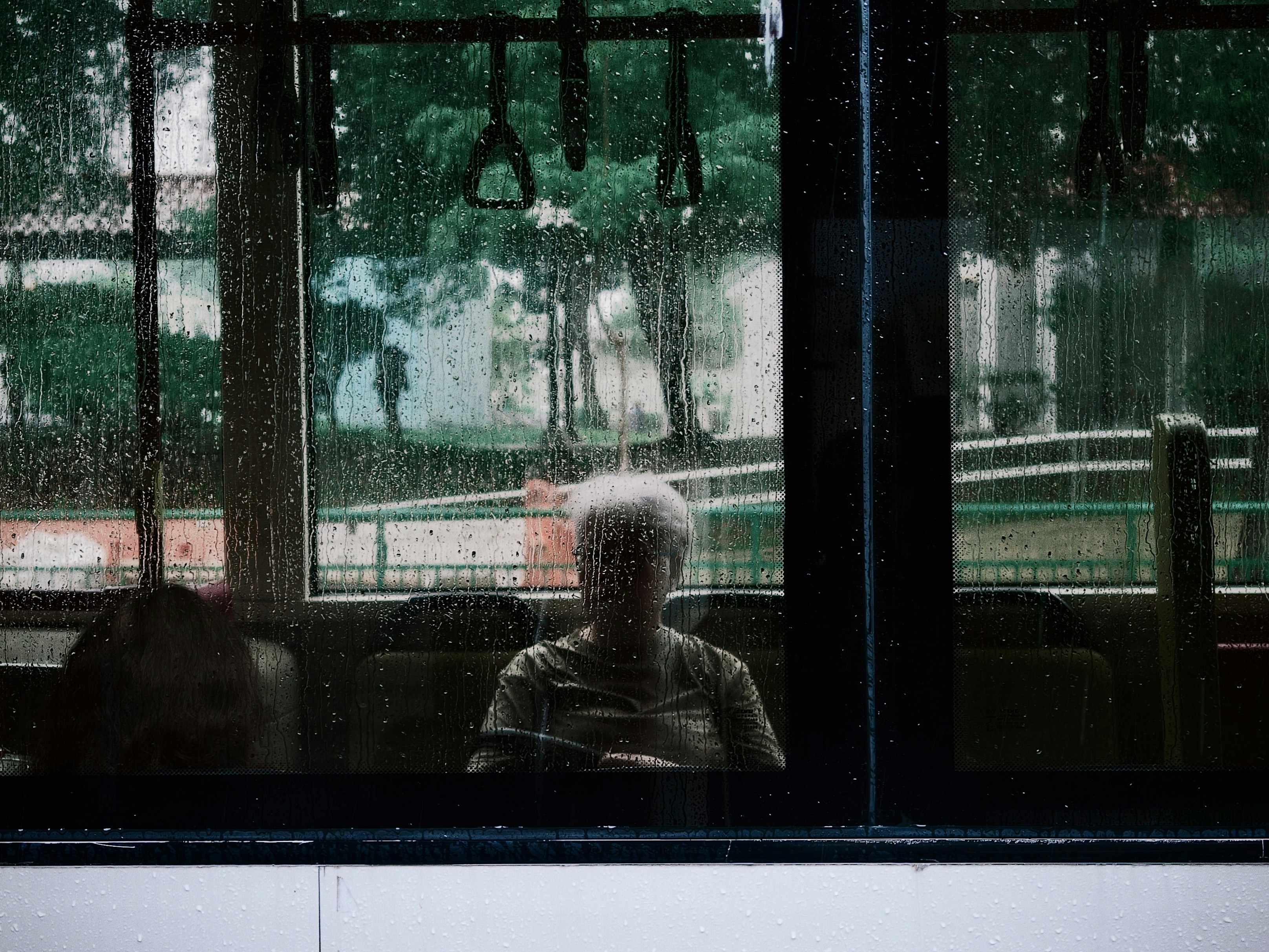 A person with grey hair seen through a rain-streaked bus window