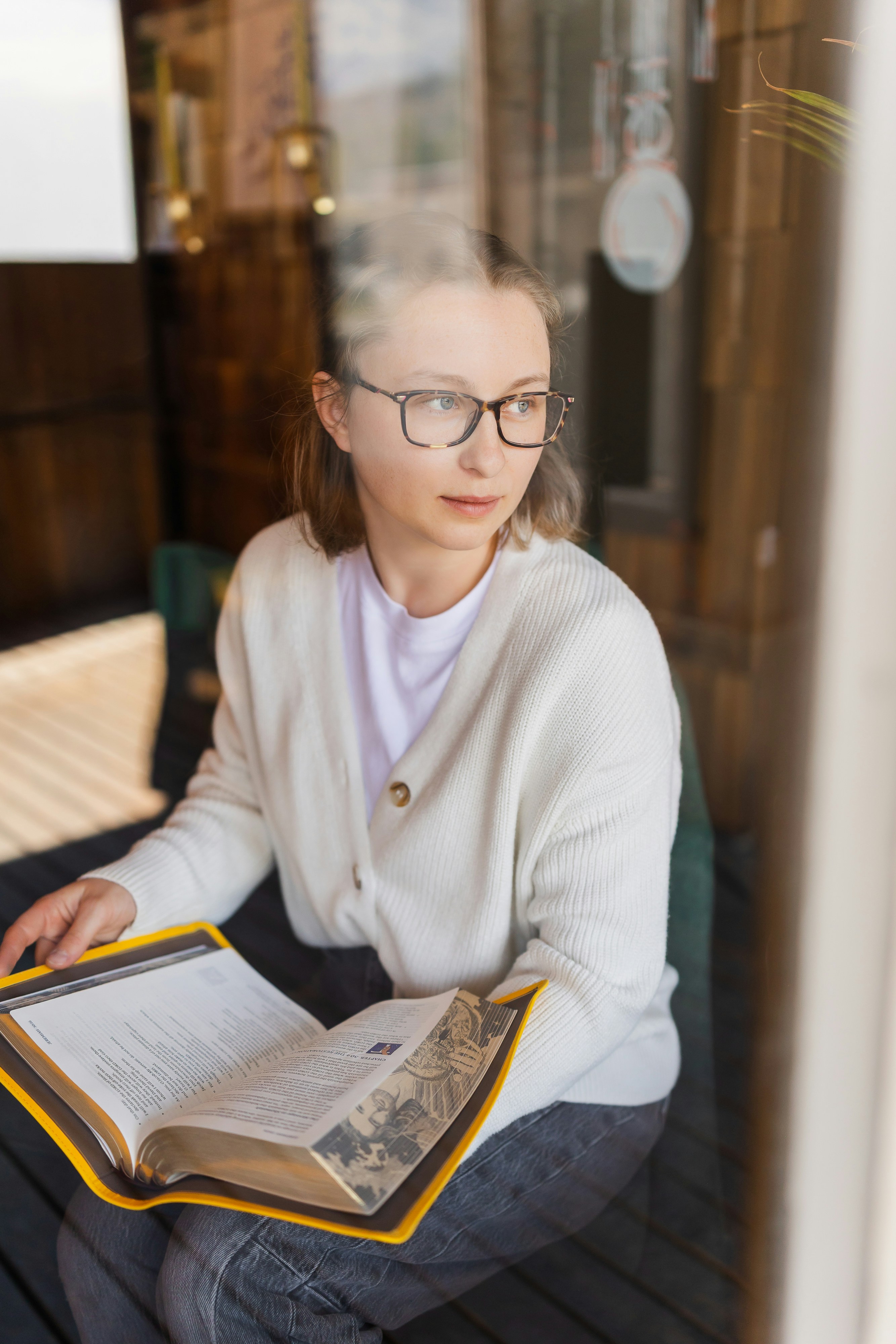 Una donna con cardigan bianco e occhiali che legge un libro in un caffè