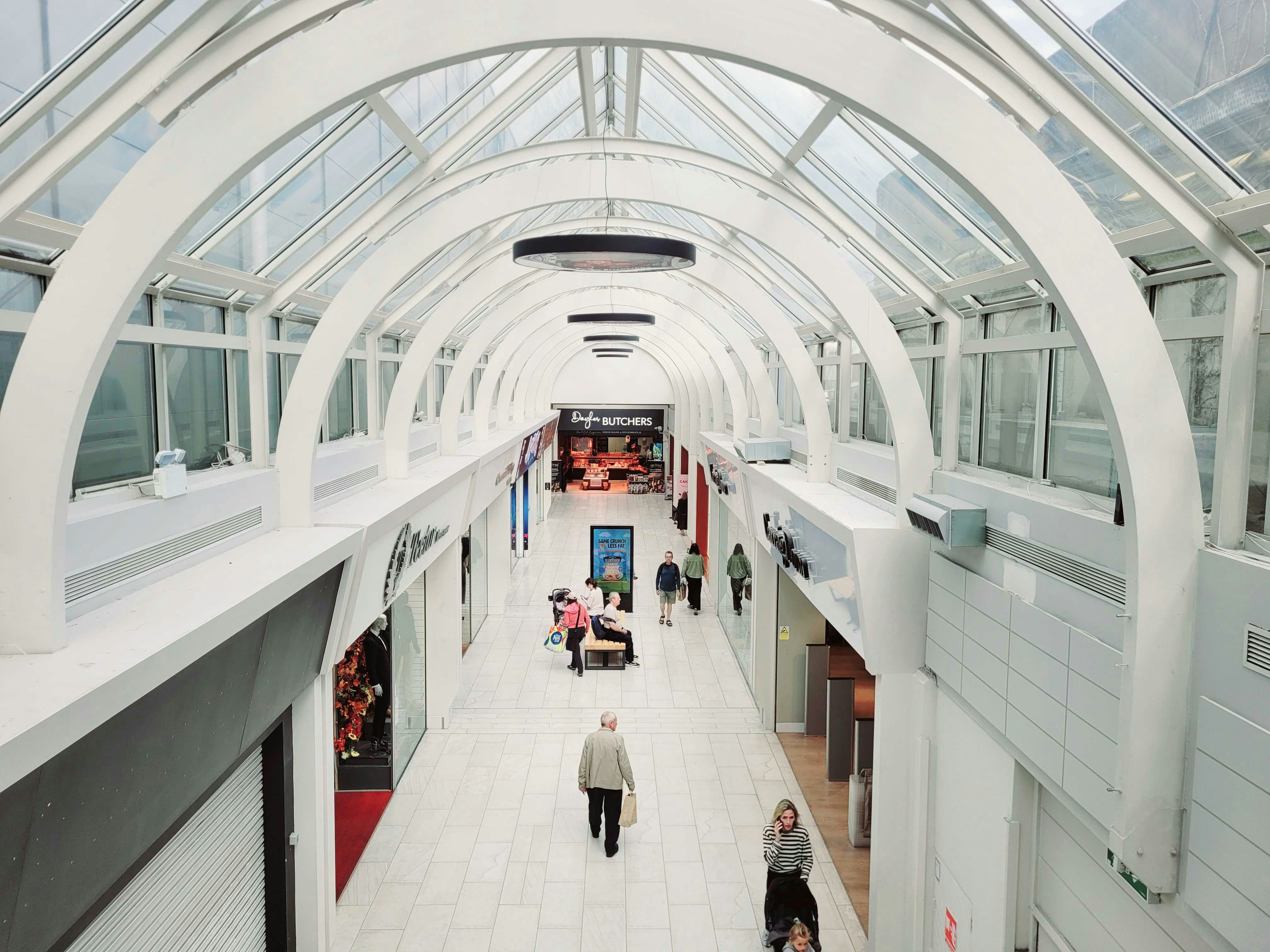 Orang-orang berjalan melewati arcade perbelanjaan yang terang dengan langit-langit kaca lengkung tinggi