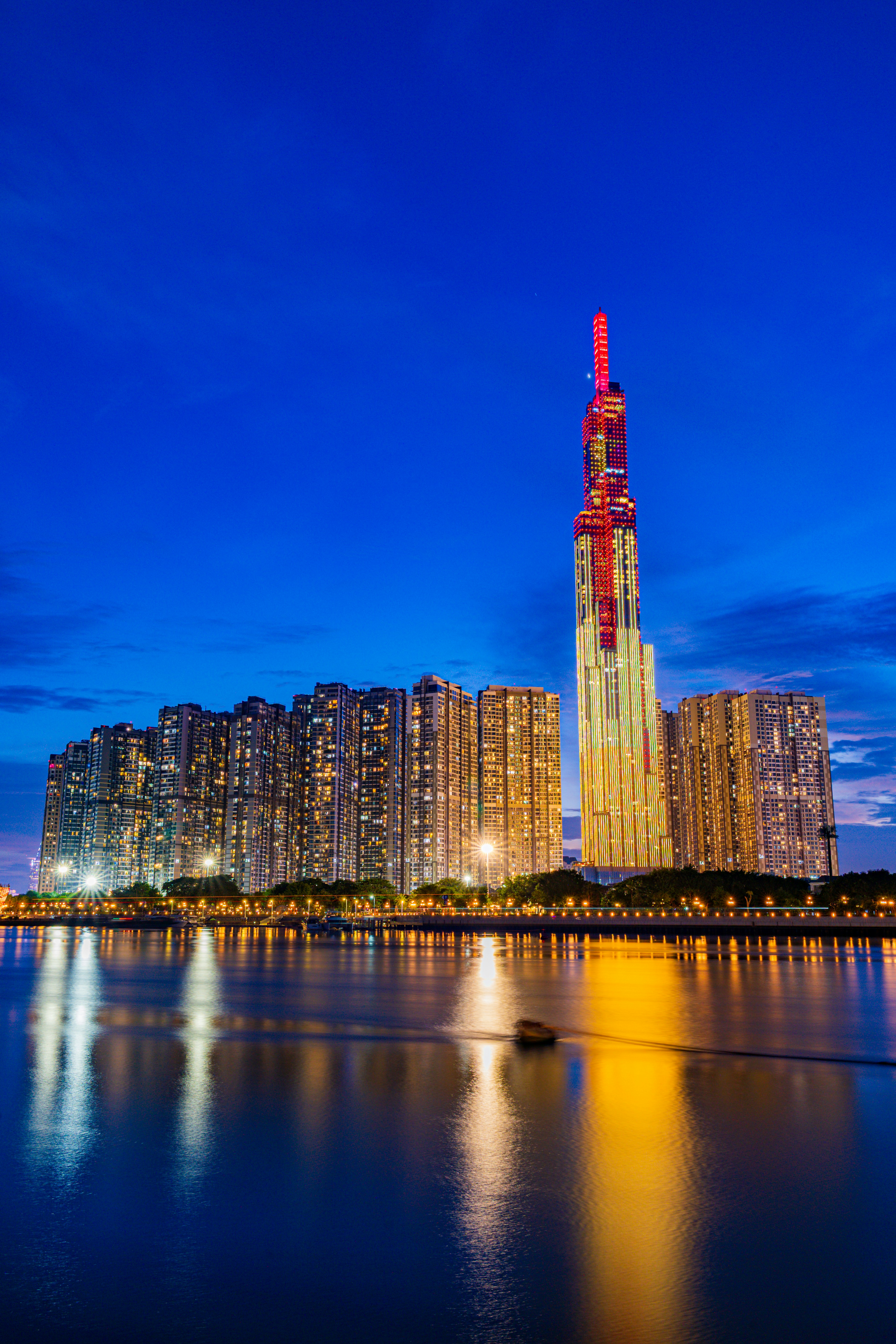 A tall illuminated skyscraper and surrounding apartment buildings reflected in a river at night