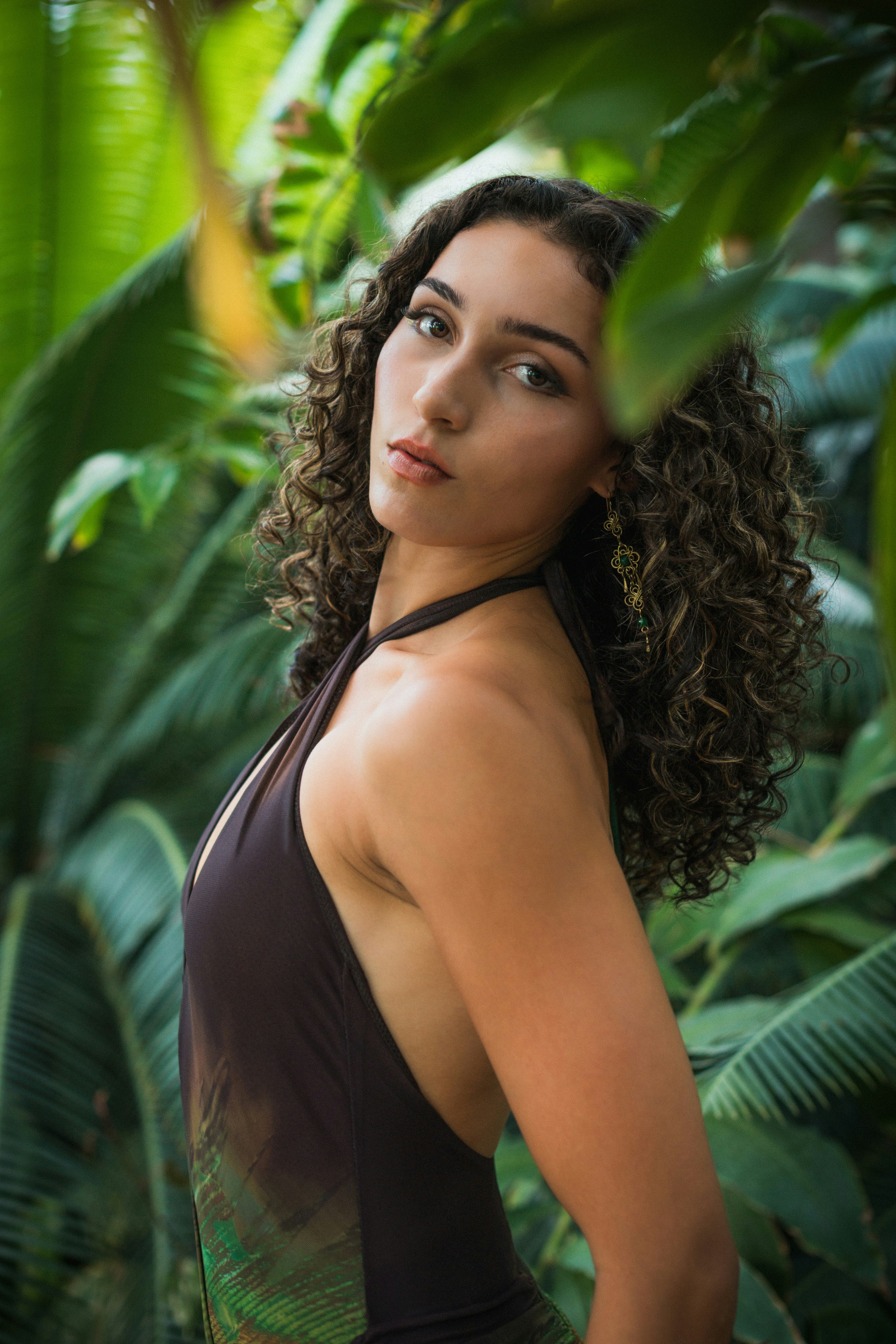 A woman with curly hair wearing a dark halter top among green palm leaves