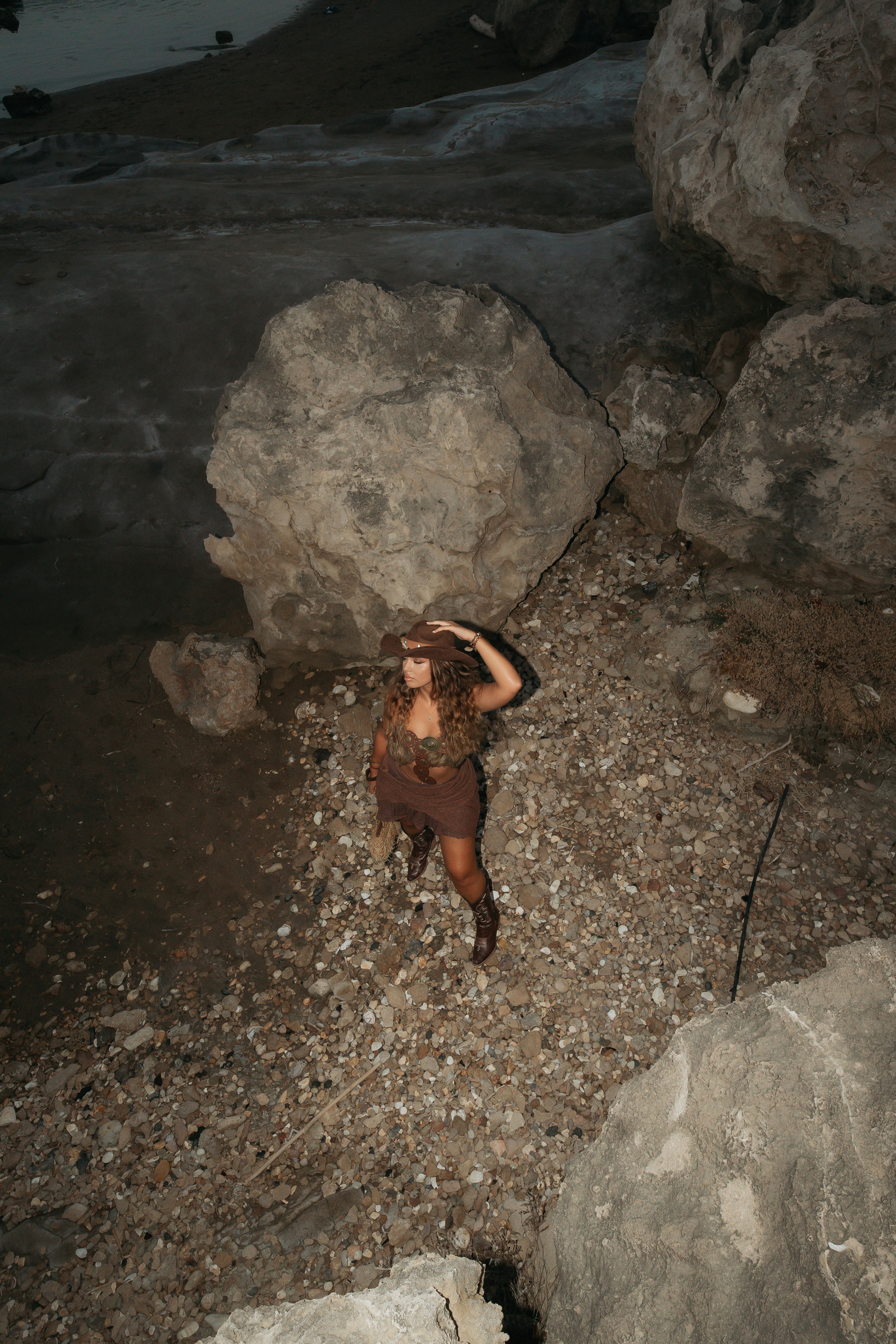 A woman in a brown cowboy hat and boots standing on a rocky beach