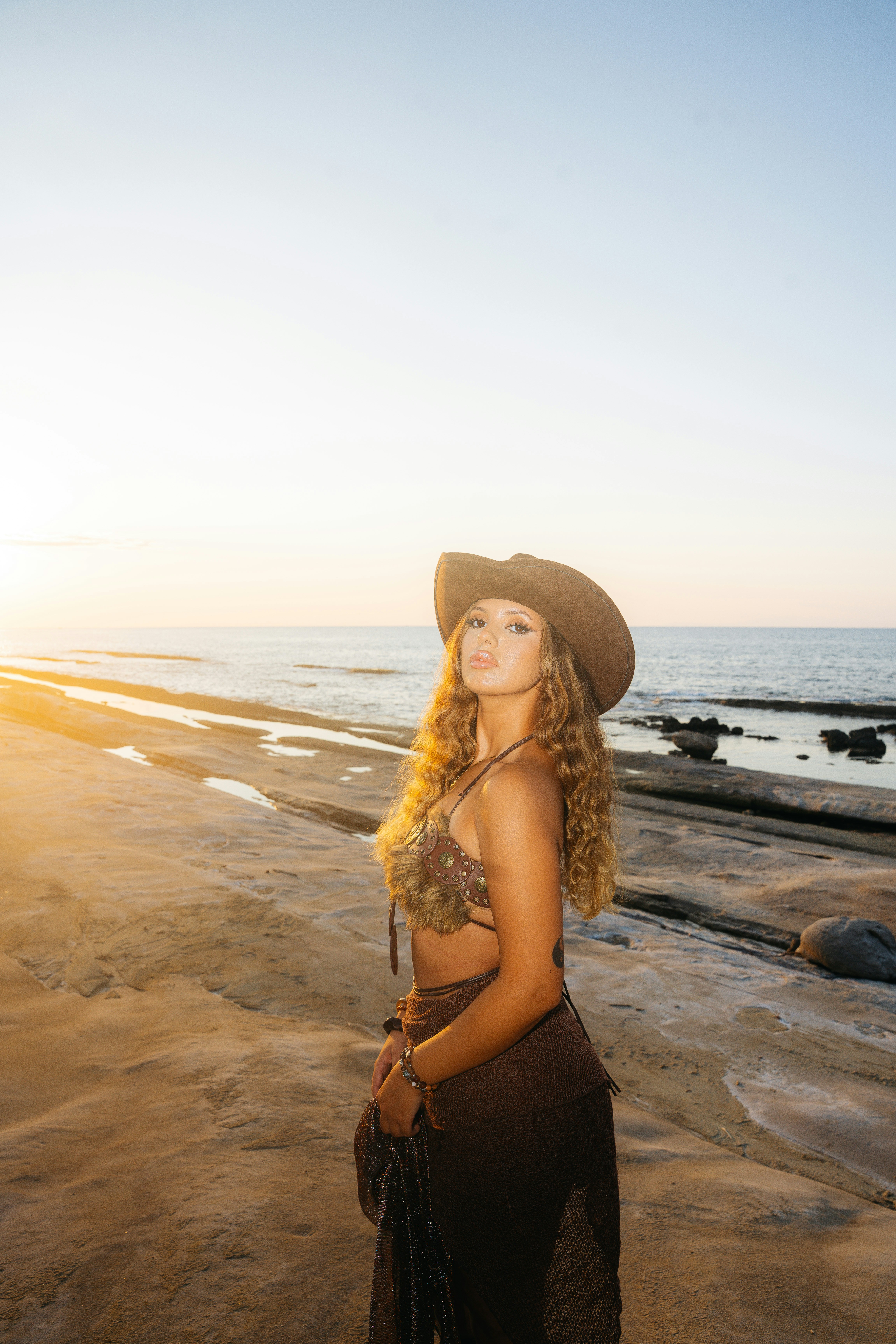 A woman in a brown hat and top on a sandy beach at low sun