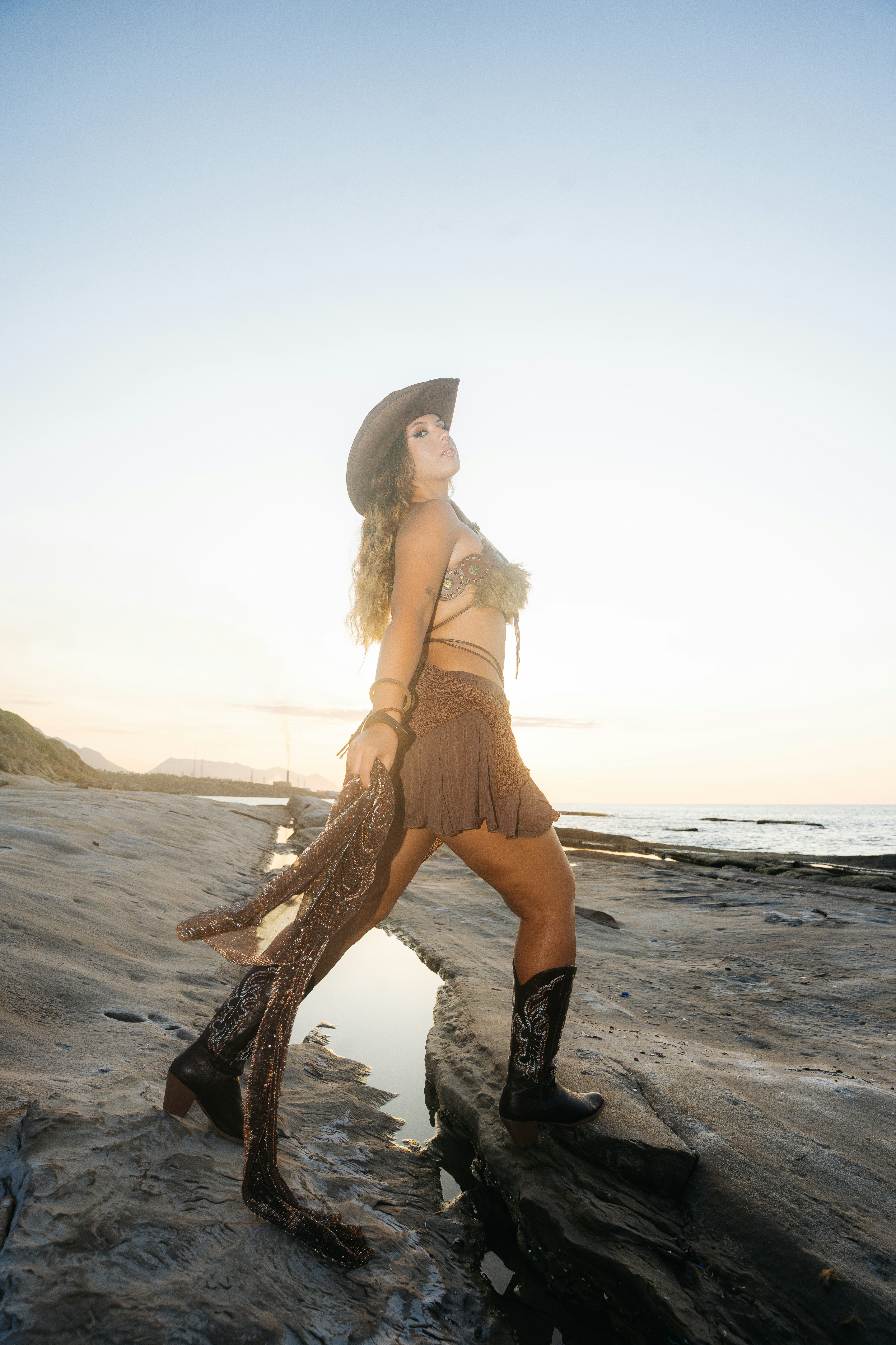 A woman in a cowboy hat and boots on a rocky beach at golden hour