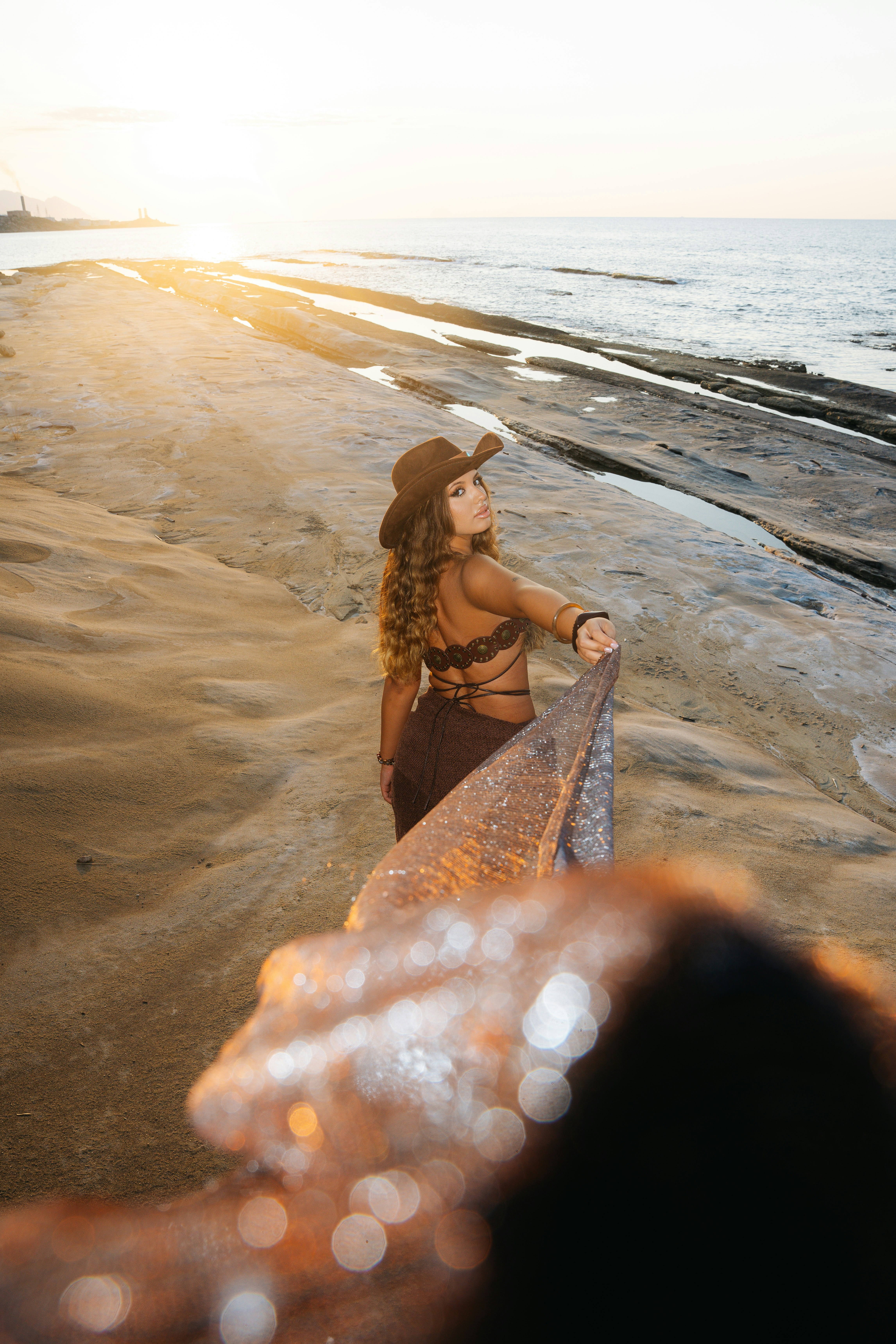 A woman in a brown hat and top holding a shimmering fabric on a beach