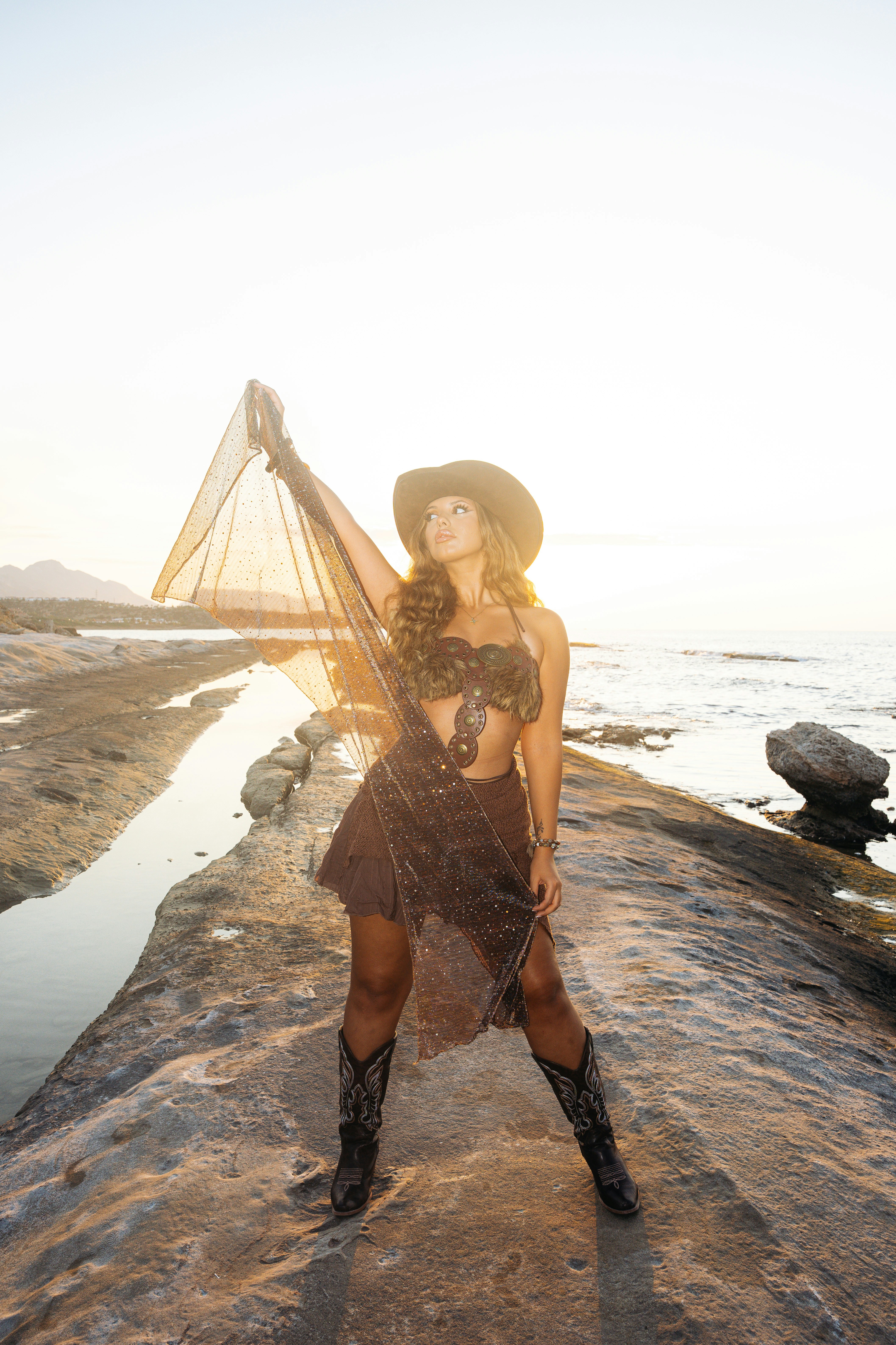 A woman holding a sheer fabric on a rocky shore at golden hour