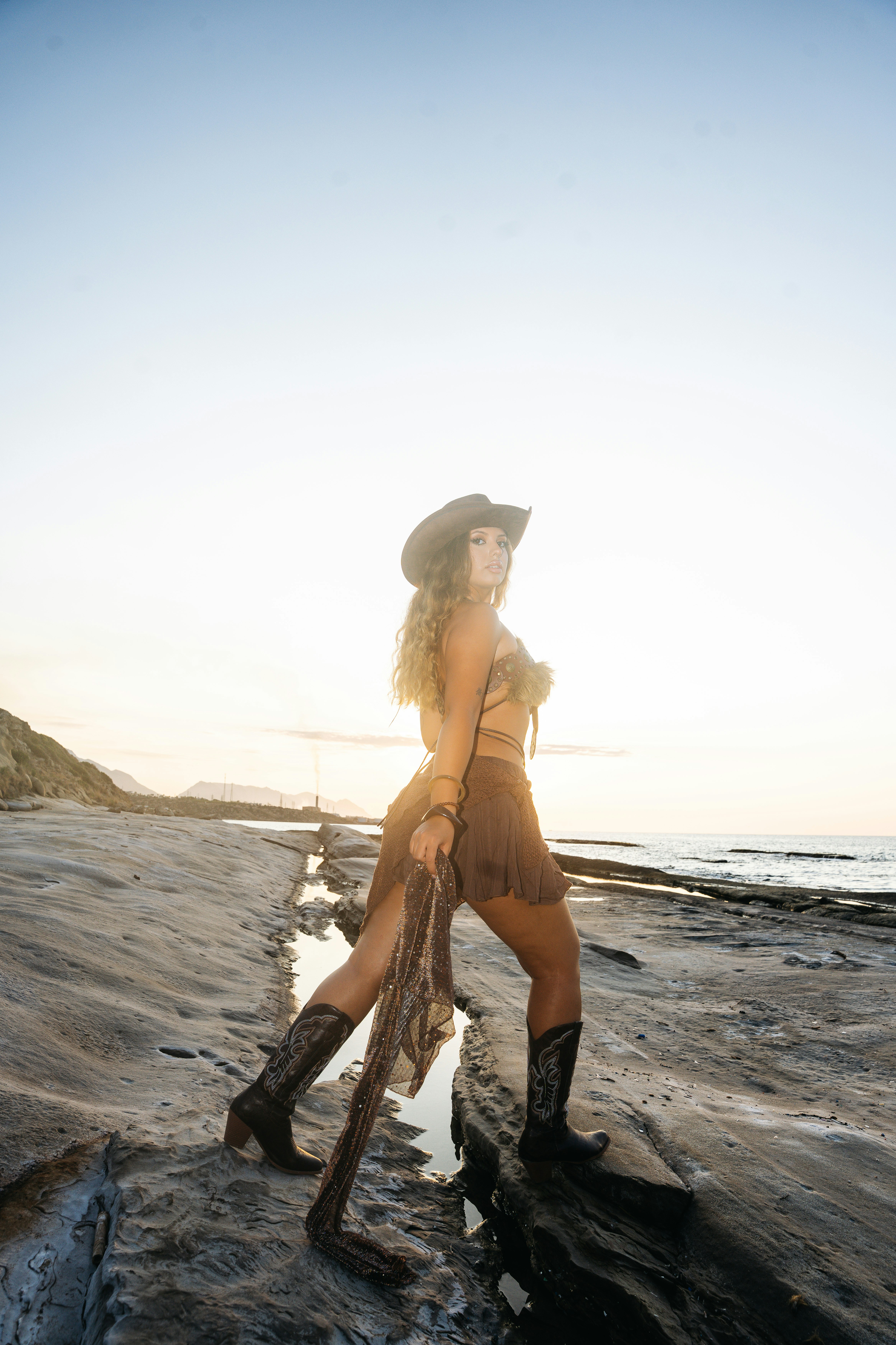 A woman in a cowboy hat and boots on a rocky shore at golden hour