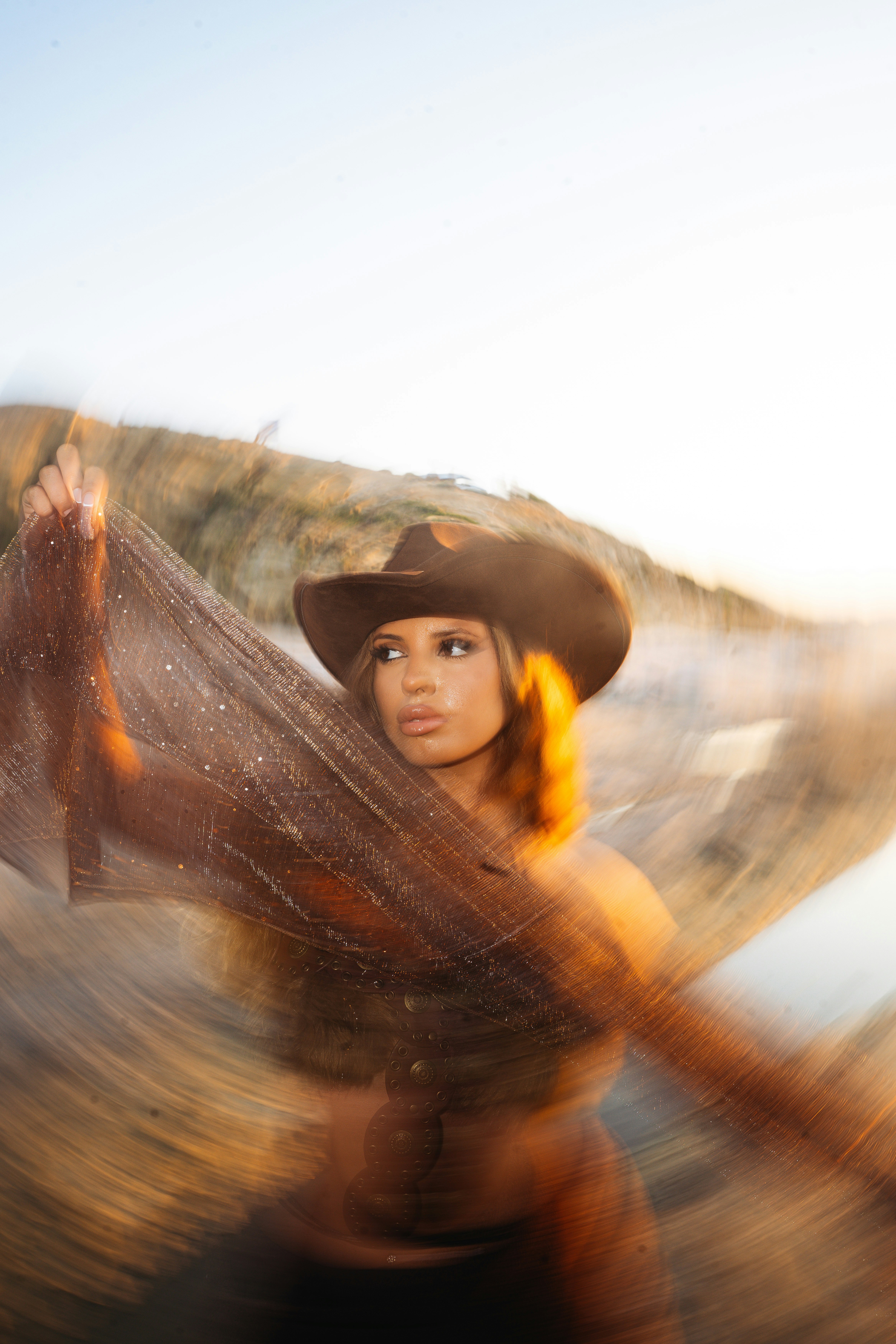 A woman in a brown cowboy hat holding a shimmering fabric in golden light