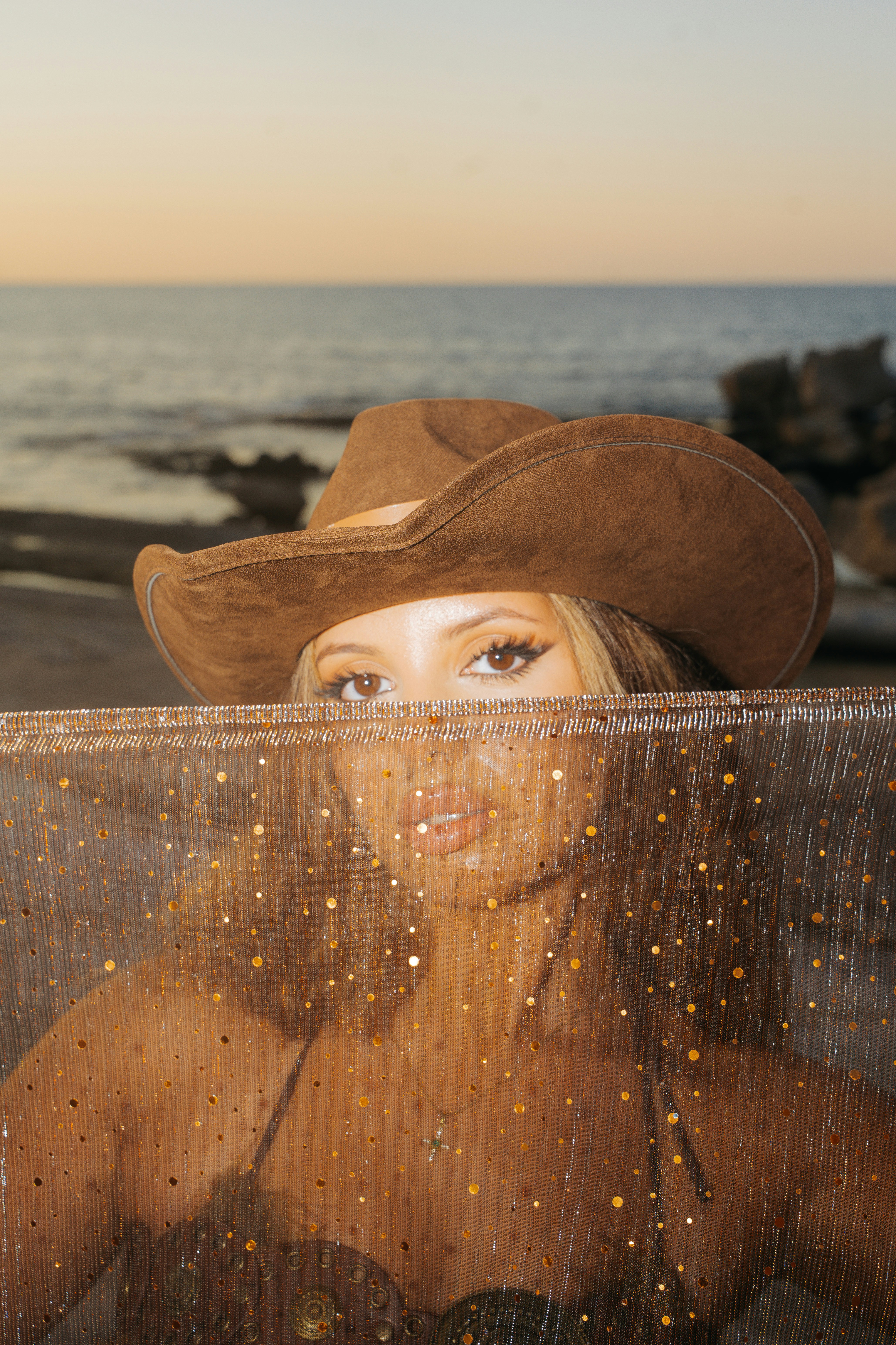 A woman in a brown cowboy hat behind a shimmering gold veil at the beach