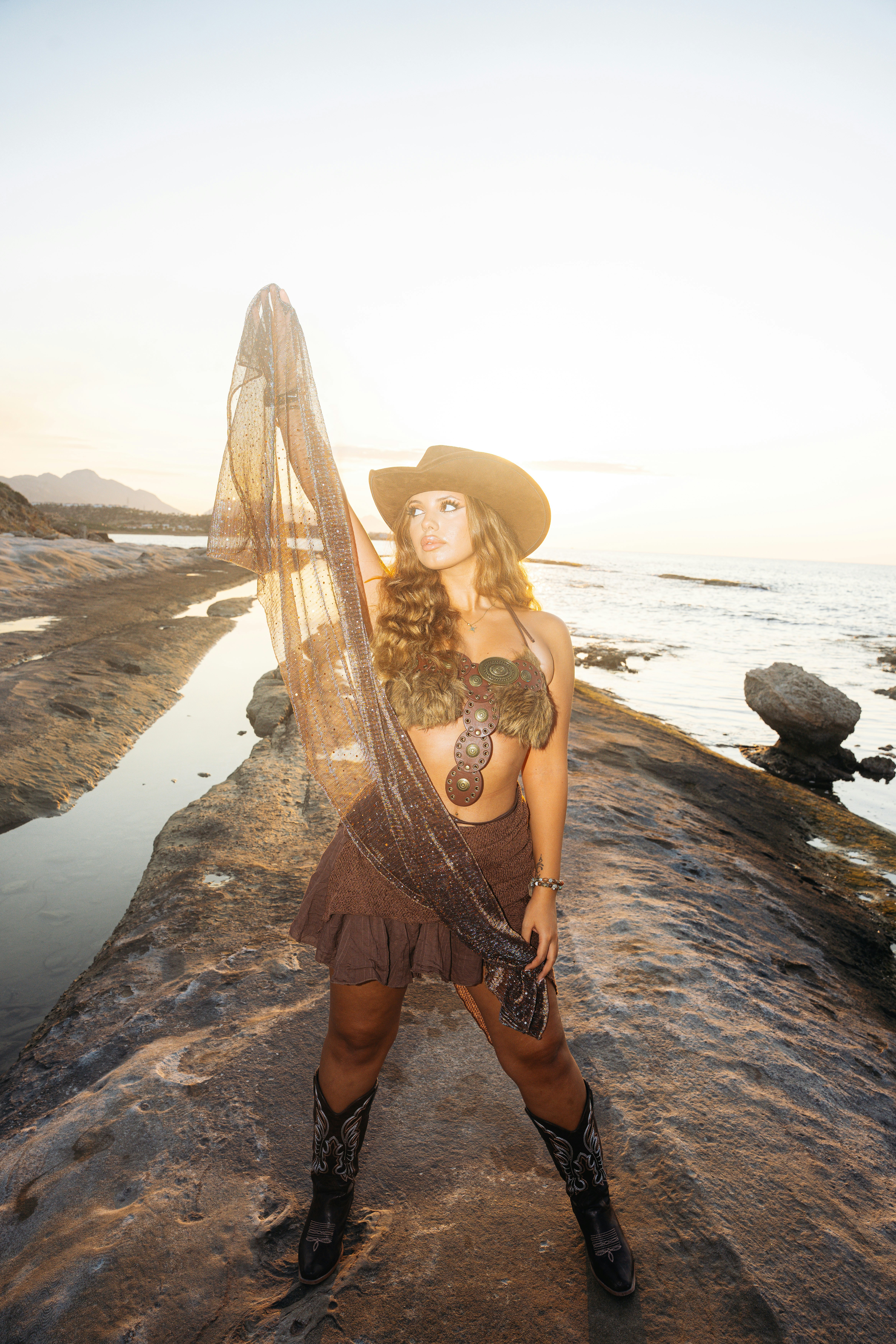 A woman holding up a sheer scarf on a rocky coastal shore at golden hour