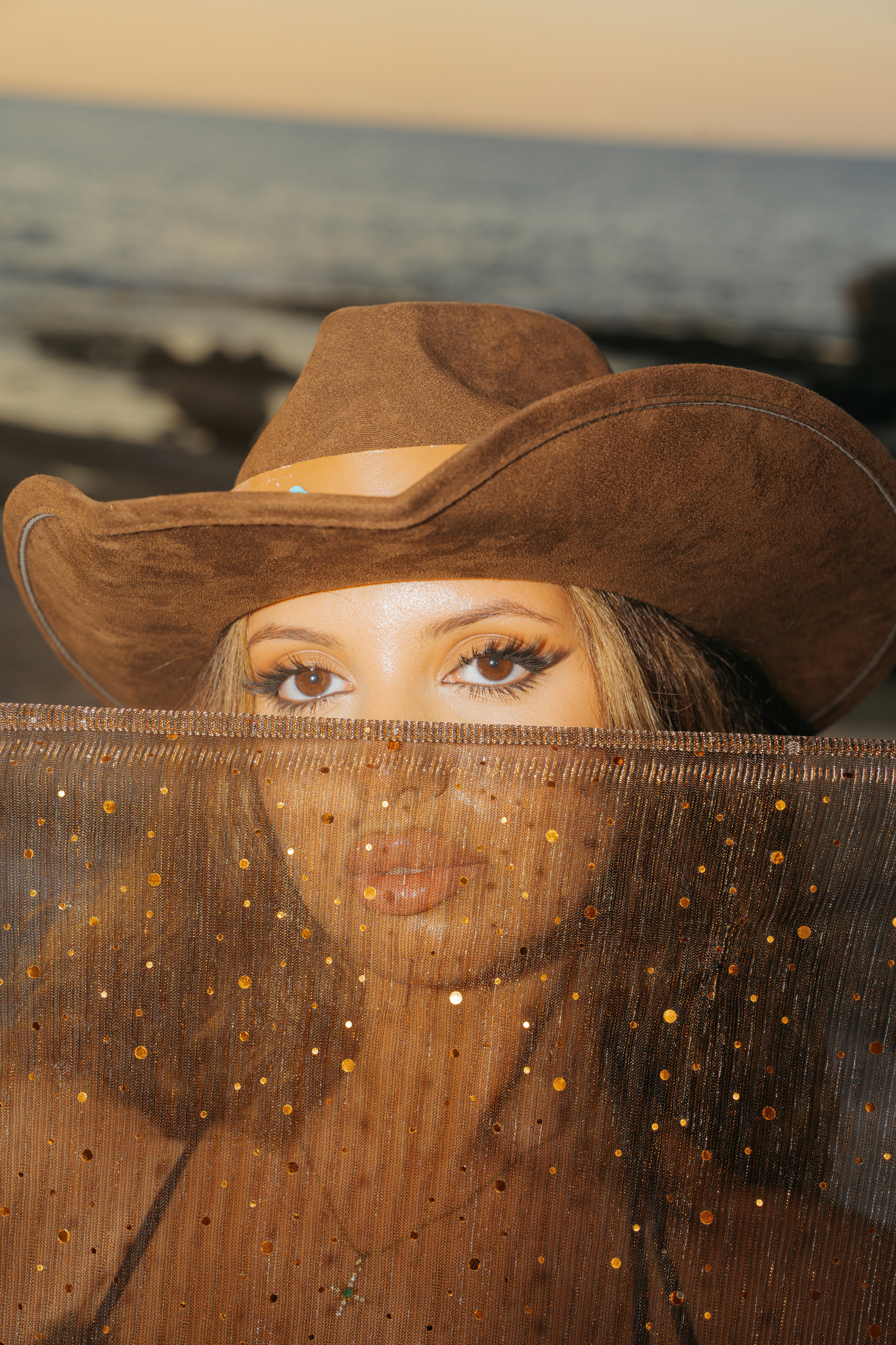 A woman in a brown cowboy hat holding a sparkling veil over her face