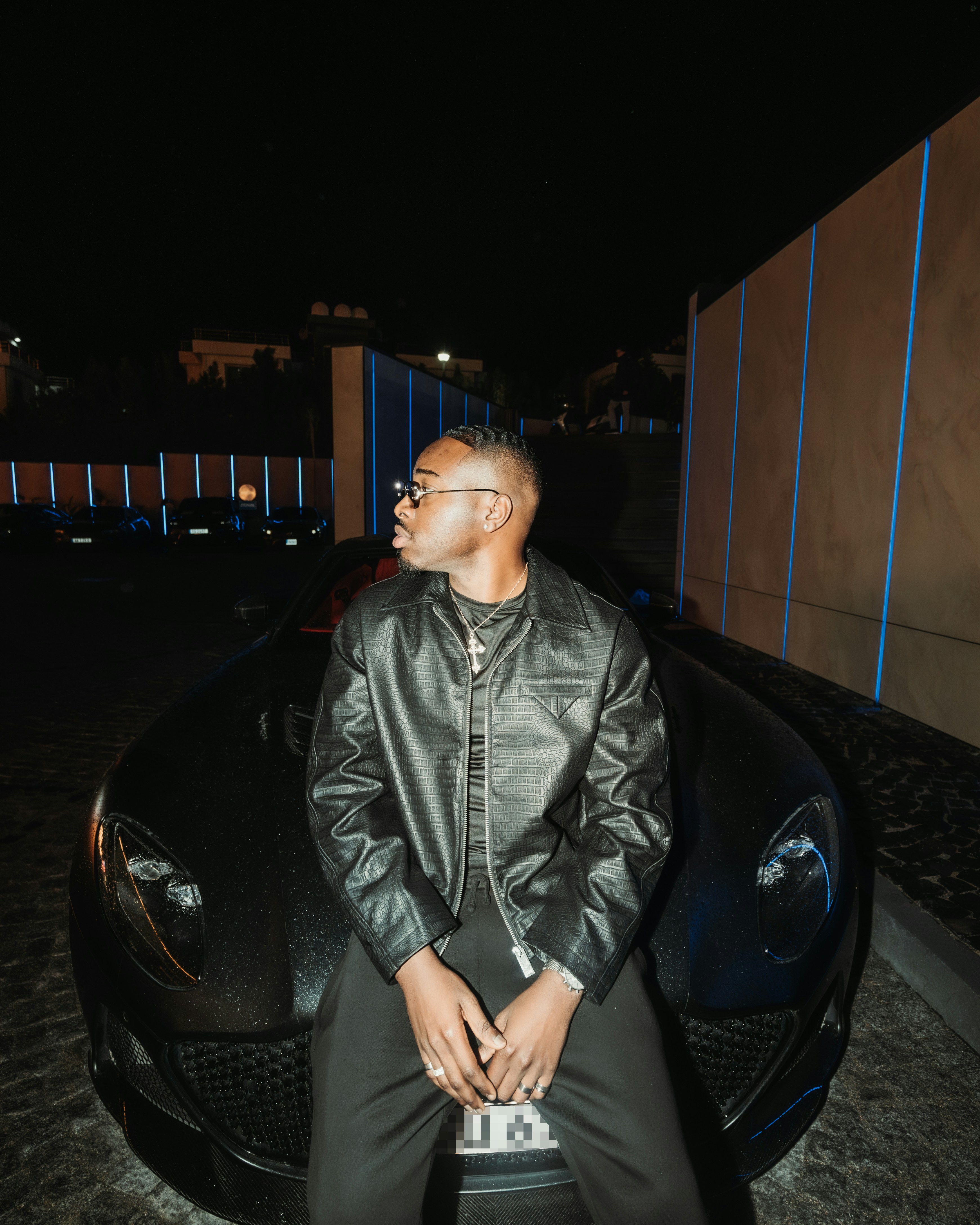 A man in a black textured jacket sitting on a dark sports car at night